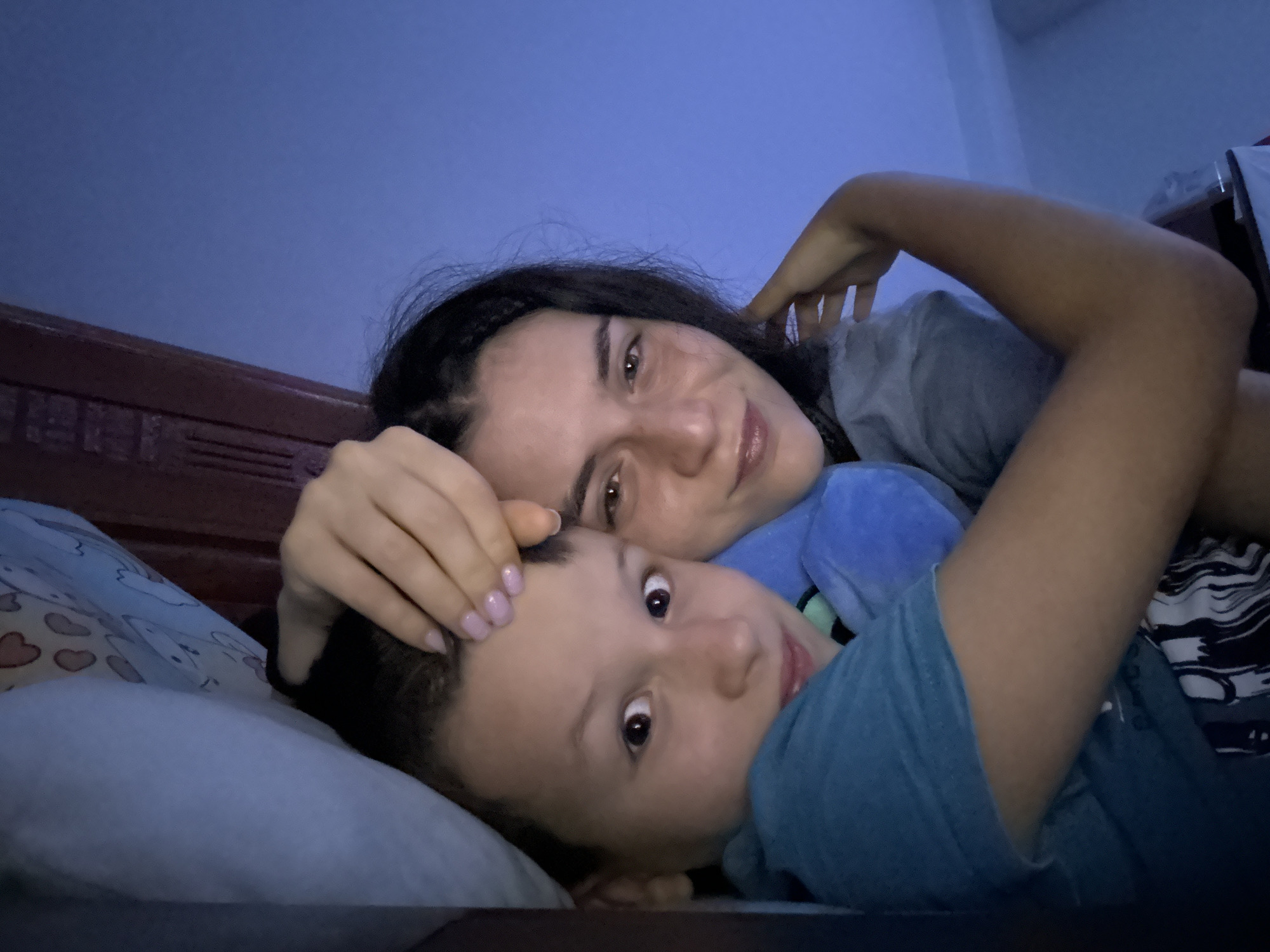 A woman and a child are lying in bed. The woman has dark hair and light skin, and is wearing a gray t-shirt. She is resting her hand on the child's forehead. The child has short hair, and is wearing a blue t-shirt. They are both looking at the camera. The background is a bedroom, with a blue wall and a wooden headboard. The location is Mũi Né, Vietnam.