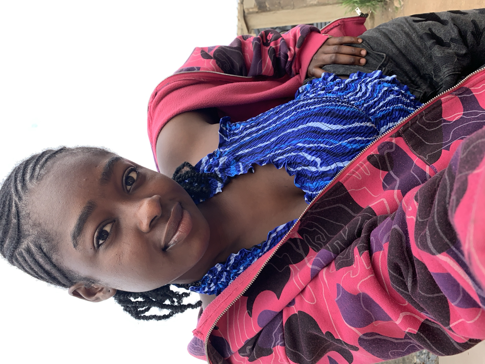 A young woman with dark skin and braided hair looks directly at the camera, smiling faintly. She is positioned for a selfie, with the camera angled upwards from a low vantage point. She wears a blue and white smocked top with ruffled details, partially covered by an unzipped hoodie featuring a pink, purple, and black camouflage pattern. Her left hand rests on her dark grey or black pants. The background is overexposed white, possibly a bright sky. To the far right, a light-colored building and hints of uneven ground suggest an outdoor, possibly semi-urban or rural, environment in Rafin Par, Nigeria.