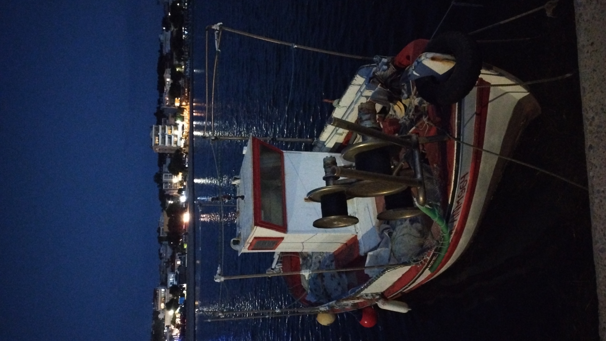 A fishing boat is docked in the dark waters of Neoi Epivates, Greece, at dusk. The boat, painted white with red trim, is equipped with two large brass winches for fishing nets. The hull of the boat bears the registration number "ANM196H". In the background, the city lights of Neoi Epivates illuminate the shoreline, with several buildings and trees visible. The sky is a deep blue, indicating the transition from day to night. The water is calm, reflecting the distant lights. A tire fender is attached to the side of the boat. There are no people visible in the image.