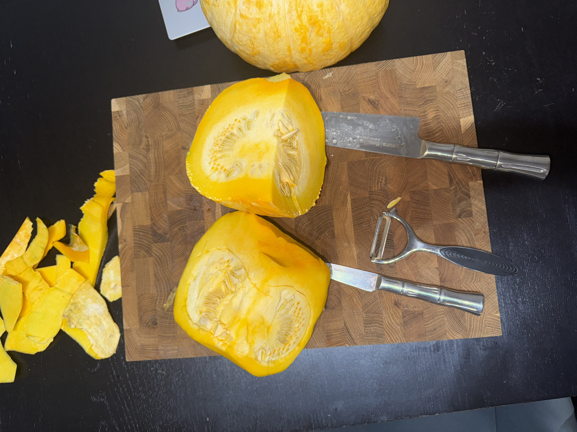 A partially cut yellow pumpkin is positioned on a wooden cutting board. Two halves of the pumpkin, showing seeds and pulp, are laid flat. Next to them are a paring knife and a peeler. The cutting board is placed on a dark, textured surface, likely a table. Above the board, scattered pumpkin peels are visible. A whole pumpkin is partially visible on the right side of the image. The scene suggests food preparation. The location is not specified in the image or provided information, therefore it cannot be determined to be in the city of Perm, Russia.