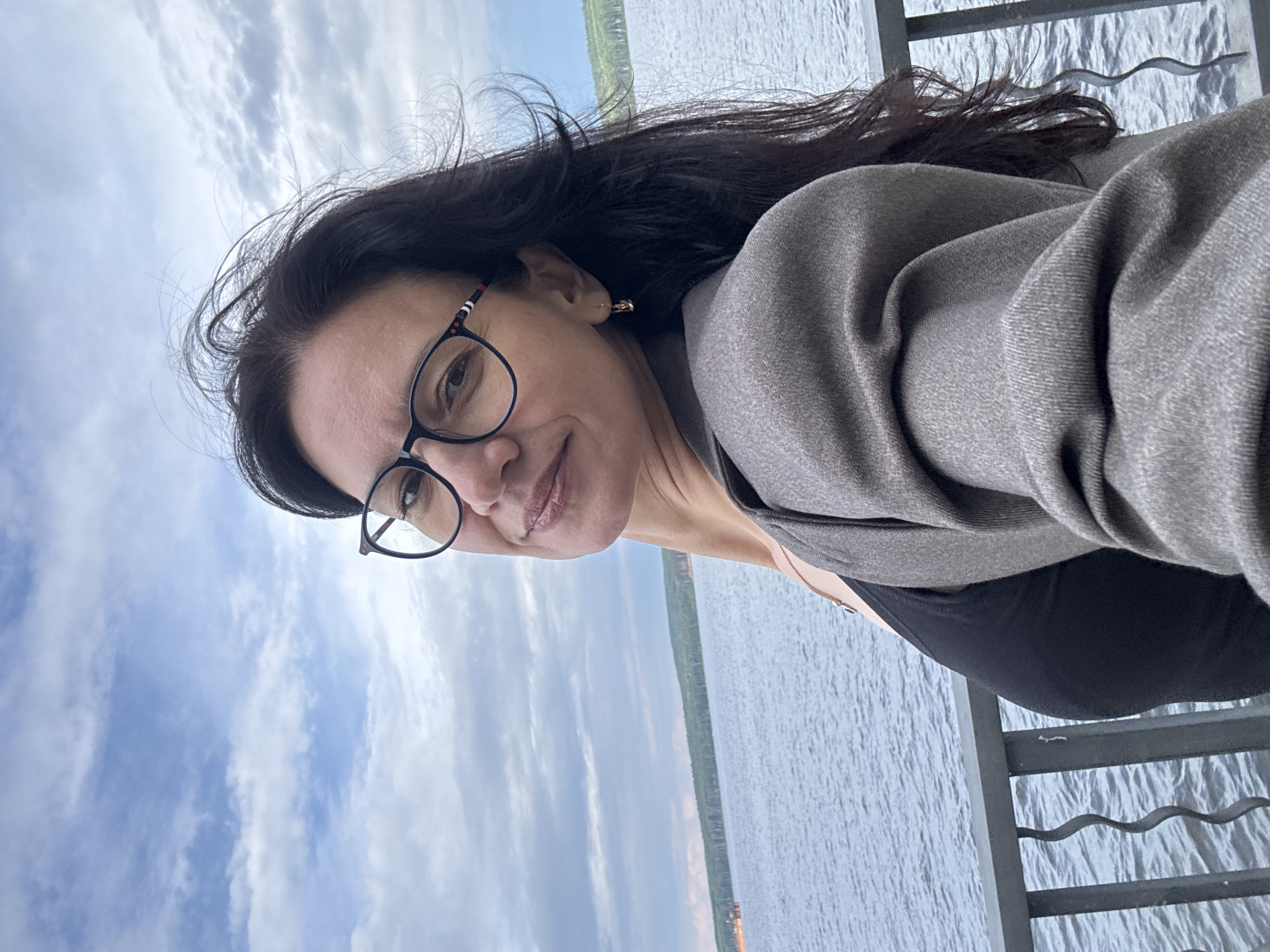 A medium shot photograph captures a woman with dark hair and glasses posing for a selfie outdoors. She is positioned in the foreground, with a body of water and a treeline in the background. The woman is wearing a gray jacket over a black top and a delicate necklace. Her hair is blowing in the wind. The scene appears to be a waterfront area, possibly a promenade or embankment, given the presence of a railing. The sky above is cloudy. The location is the city of Chaikovsky, Russia.