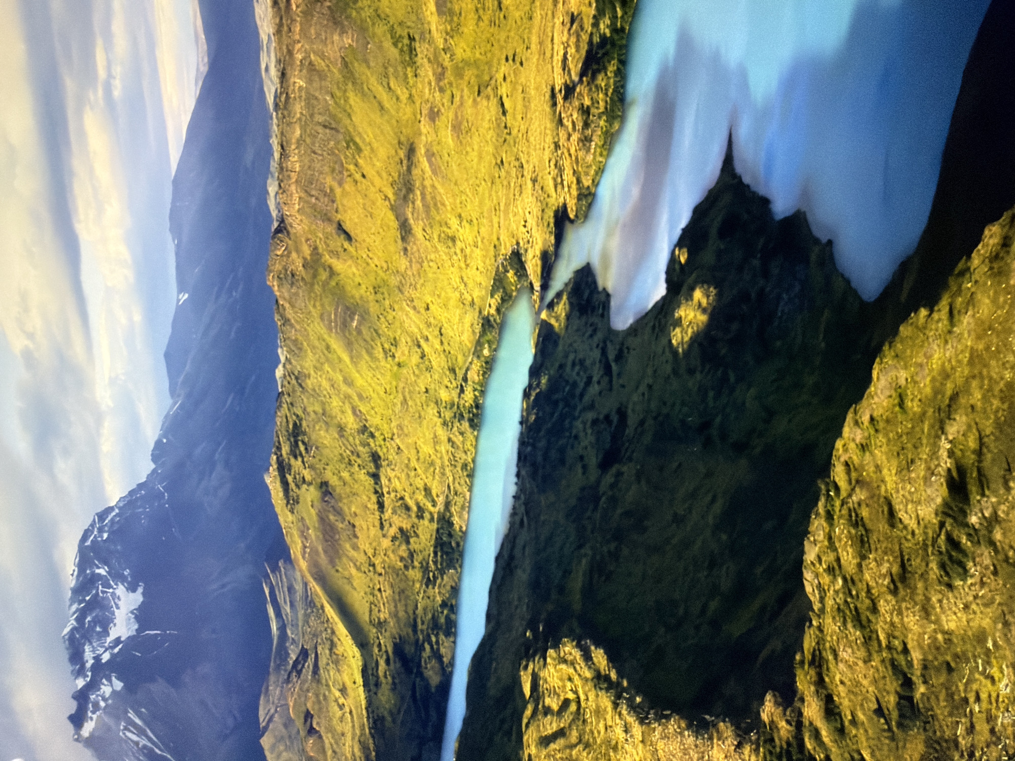 The image depicts a dramatic mountain landscape under a cloudy sky. In the foreground, a bright turquoise river or lake winds through rolling hills covered in vibrant green and yellow vegetation. The terrain is rugged, with steep slopes and shadowed ravines. In the background, majestic mountains rise, their peaks capped with snow, suggesting a high-altitude or polar environment. The lighting is soft, creating a serene yet powerful atmosphere. This landscape is not located in Kyiv, Ukraine; it is a natural mountain and river scene.