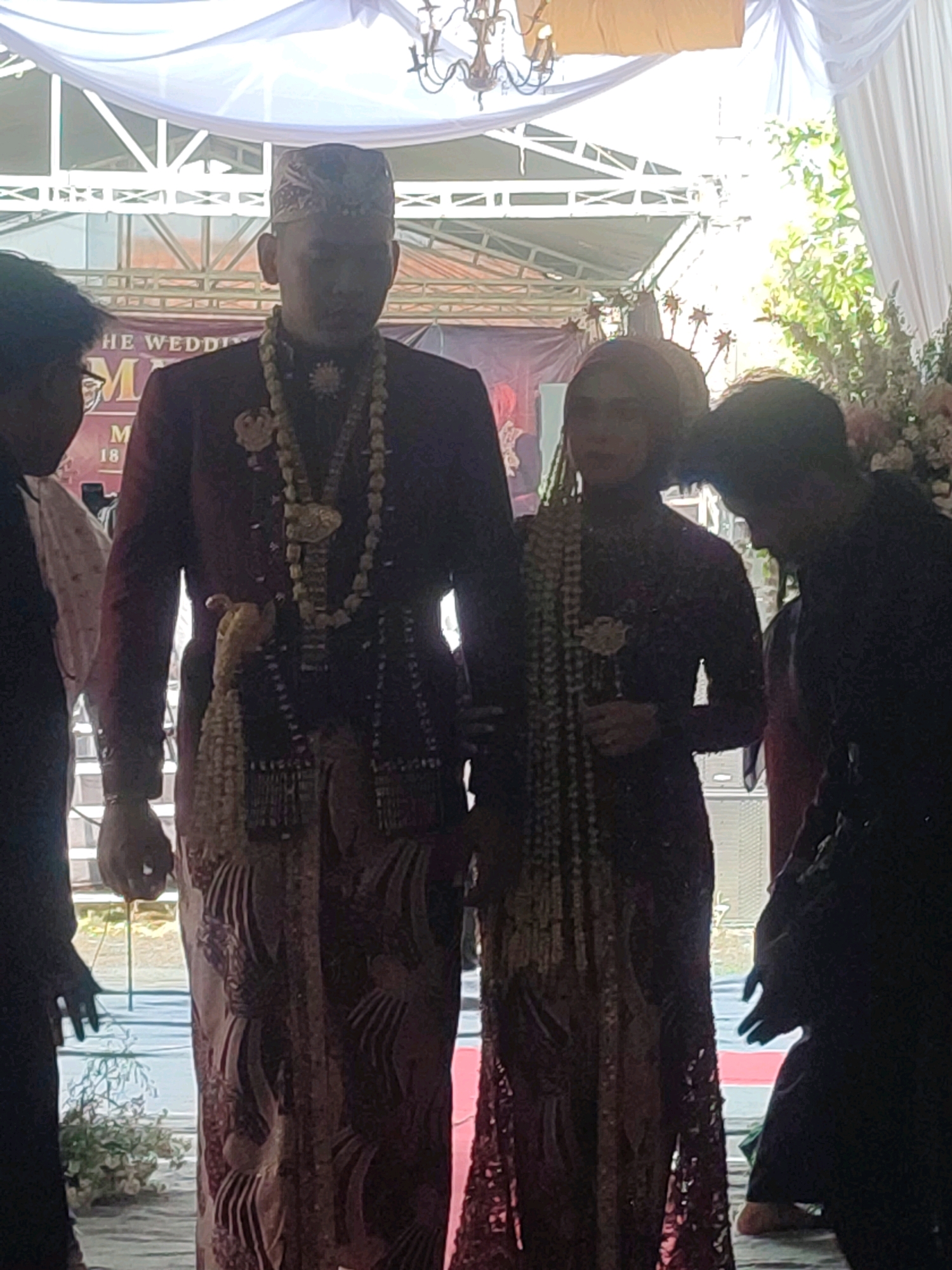 A wedding ceremony is taking place in an outdoor setting, likely in Kecamatan Haurgeulis, Indonesia, as indicated by a banner in the background that reads "THE WEDDING M 18". The central figures are a bride and groom, dressed in traditional wedding attire. The groom wears a dark maroon jacket with intricate gold embroidery and a matching patterned sarong. He is adorned with multiple layers of beaded necklaces and a decorative sash. The bride wears a dark patterned dress with a matching headscarf and also wears beaded necklaces. They are positioned in the center of the frame, with other wedding guests partially visible on either side, some in silhouette due to lighting. The background features a white draped tent, structural elements of a tent or canopy, and greenery from trees. The lighting suggests it is daytime, with a bright light source coming from above and behind the couple.