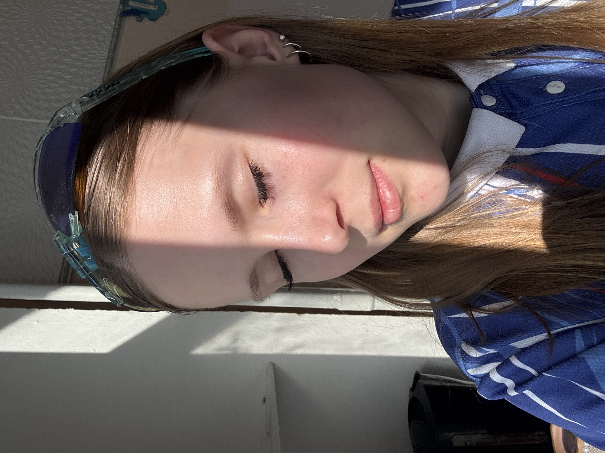 A close-up, rotated image shows a young woman's face in bright sunlight. She has fair skin, brown hair, and is wearing blue glasses on her head. She has multiple piercings in her left ear. She's wearing a blue and white collared shirt. The background shows a textured wall. The image appears to have been taken indoors. There is no verifiable information to suggest this photo was taken in Berezniki, Russia.