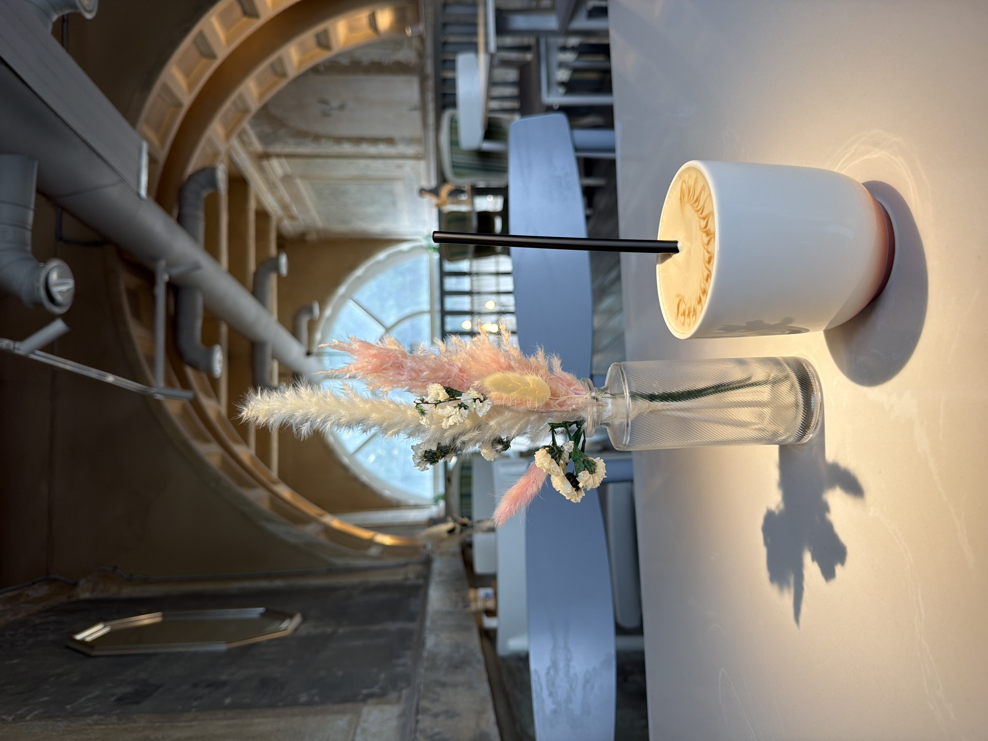 This image shows a bright, modernly decorated interior, likely a cafe or restaurant, in Moscow, Russia. The perspective is unique, almost an aerial shot from a table. Dominating the foreground on the right is a white coffee cup with latte art and a black straw, next to a glass vase holding decorative floral arrangements in pastel colors. These sit on a clean, light-colored table.
The background is where the Moscow element comes in; the architectural style strongly suggests the city. A large arched window is visible, revealing a cityscape. Overhead, industrial-style metal pipes run along the ceiling. A golden tray sits on the surface in the lower-left, contributing to the scene's overall ambiance. The image is well-lit, with soft shadows accentuating the details of the flowers and vase.