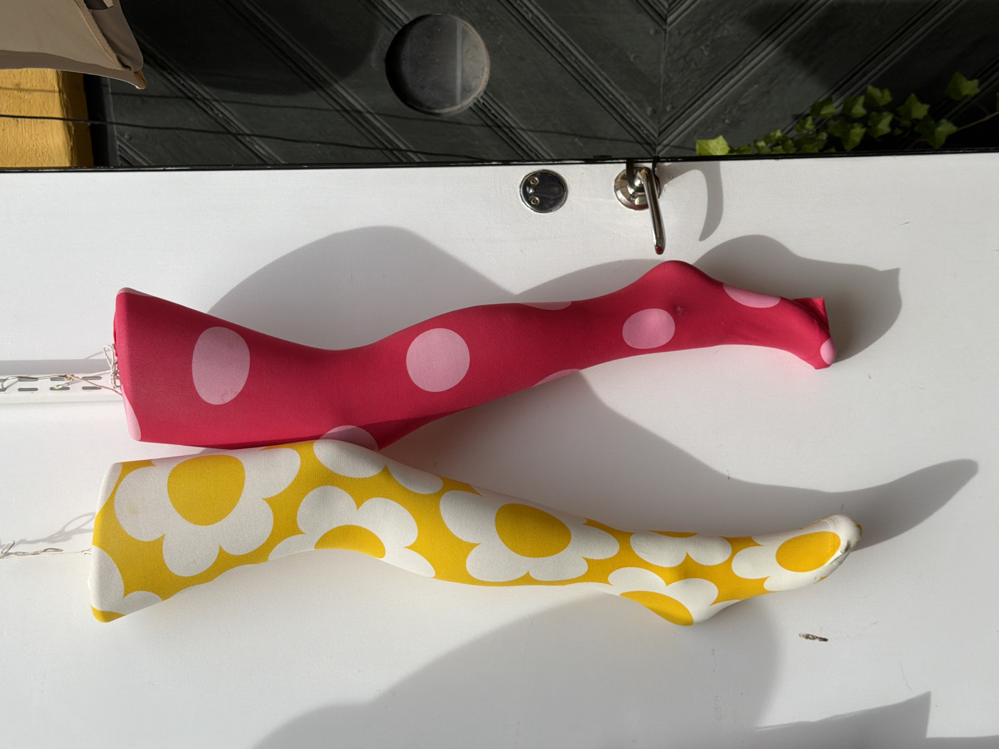 The image captures two mannequins shaped as legs, adorned with brightly colored stockings. The left mannequin is dressed in yellow stockings featuring a bold white floral pattern, while the right mannequin sports pink stockings with white polka dots. These mannequins are suspended by wires against a white wall. To the right of the wall, a dark, paneled door is visible, equipped with a silver doorknob and a circular window. A hint of green ivy grows along the edge of the door. The scene appears to be an outdoor display, possibly in Malmö, Sweden, given the architectural style of the door and the general aesthetic. The lighting suggests it is daytime, with shadows cast by the mannequins and the door.