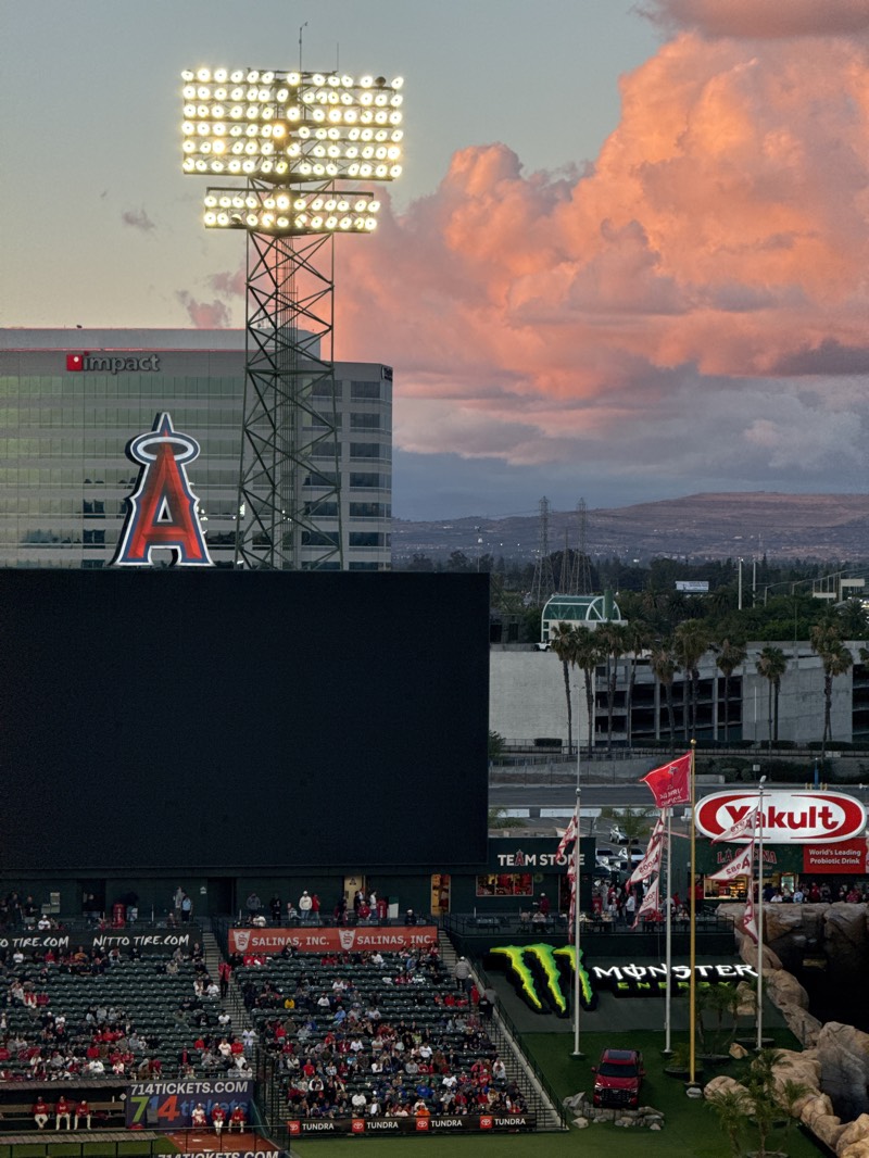The image depicts a vibrant scene at an outdoor baseball stadium during sunset or dusk in Anaheim, United States. In the foreground, numerous spectators are seated in red stadium seats, facing a large, black scoreboard or video screen. Below the screen, sponsor banners for "NITTO TIRE.COM" and "SALINAS, INC." are visible, along with "714TICKETS.COM" on a railing.
A towering stadium light pole, brightly illuminated with multiple floodlights, stands prominently in the midground. Behind it, a modern building features the "impact" logo and a large, iconic red "A" with a halo, representing the Los Angeles Angels baseball team. To the right, a large green "MONSTER ENERGY" logo is displayed on a sloped grassy area, with a red pickup truck (likely a "TUNDRA") parked nearby. Flags, including one with the Angels logo and another for "Yakult" (which advertises "World's Leading Probiotic Drink"), fly from poles.
The sky above is a notable detail, filled with large, fluffy clouds bathed in warm pink and orange tones from the setting sun, contrasting with the cooler blue sky overhead. In the distant background, a hazy urban landscape with hills and mountains is visible. The stadium lights are fully operational, indicating the transition from day to night. The presence of the Angels logo and "714TICKETS.COM" strongly places the scene at Angel Stadium of Anaheim.