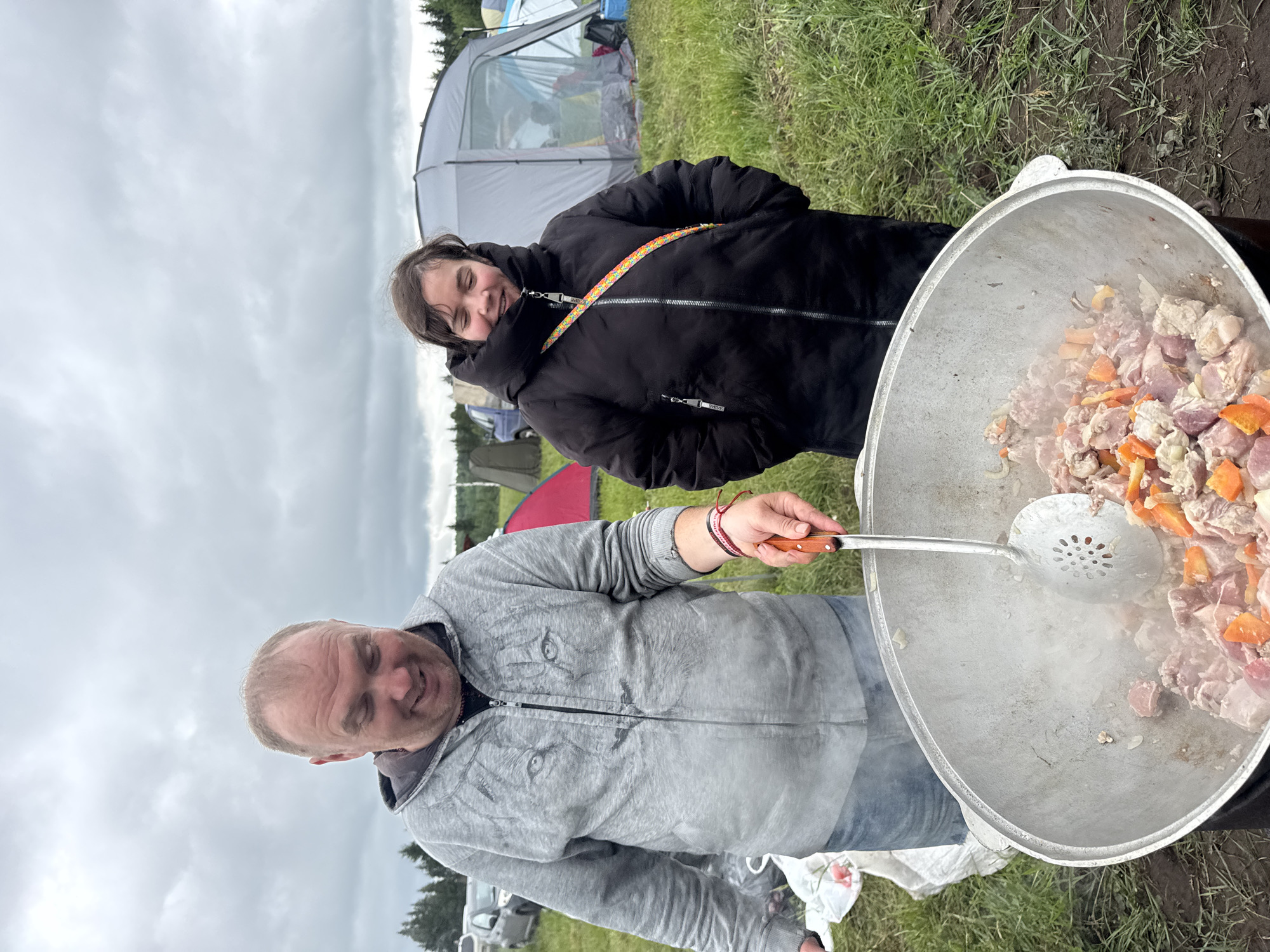 An outdoor scene in Большая Кивара, Russia, depicts two individuals engaged in large-scale cooking under an overcast sky, with a campsite visible in the background.
In the foreground, a light-skinned adult male stands on the left, smiling as he looks down into a large, silver-colored metal cooking pot. He wears a grey zippered hoodie featuring a prominent lion's face graphic and blue jeans. A red and white wristband is on his right wrist. He holds a large metal slotted spoon with a wooden handle, stirring the contents of the pot, from which steam is visibly rising. The pot contains diced meat, orange vegetables (likely carrots), and white vegetable pieces (possibly onions), actively cooking.
To the right, a light-skinned female, appearing to be a teenager or young adult, stands, also smiling and looking towards the cooking pot. She is dressed in a black puffer jacket with a high collar and a colorful patterned strap (orange, yellow, blue, white) crossing her body from her left shoulder to her right hip. Her hands are not fully visible but appear to be clasped or in her pockets.
The ground is covered in green grass, with some darker patches of dirt immediately around the cooking area. In the mid-ground and background, several camping tents are visible, including a large grey dome tent and a smaller red tent. Multiple cars are parked on the grassy terrain further to the left. A line of evergreen trees forms the distant horizon under a heavily clouded, grey sky.
