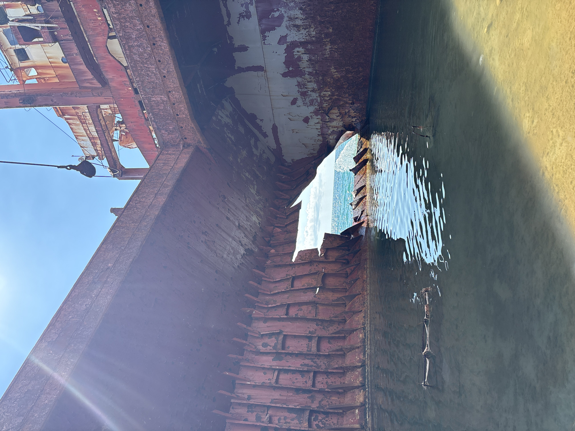 The image depicts the shipwreck of the "Sinyaya Ptitsa" (Blue Bird) in Kabardinka, Russia. The rusty hull of the vessel is partially submerged in clear, shallow water, with a sandy seabed visible in the foreground. A large opening in the hull reveals the blue sea and sky beyond. The sun is shining brightly, casting rays on the water and the weathered metal of the shipwreck. There are no people visible in the scene. The environment is one of decay and nature reclaiming a man-made structure.