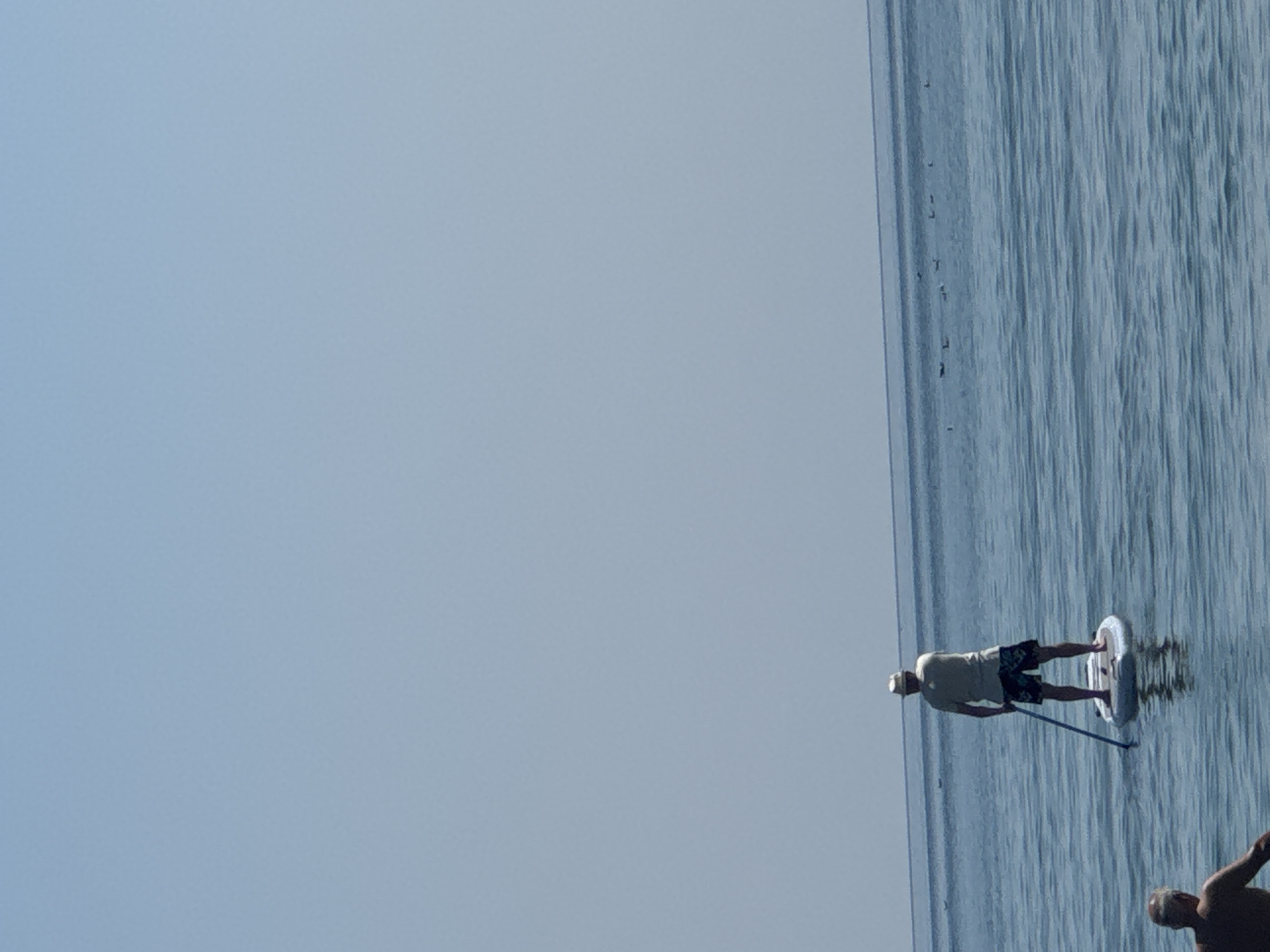 Two individuals are visible in the water. In the foreground, the back of a man with grey hair is partially visible. Further in the background, a man wearing a light-colored hat, a light-colored t-shirt, and dark patterned shorts stands on a paddleboard. He is holding a paddle. The paddleboard is floating on calm blue water. In the distance, a line of birds is visible on the water's surface. The scene appears to be set on the Black Sea coast near Sozopol, Bulgaria, indicated by the distinctive calm, clear blue water and the presence of seabirds typical of the region. The sky is clear and light blue.