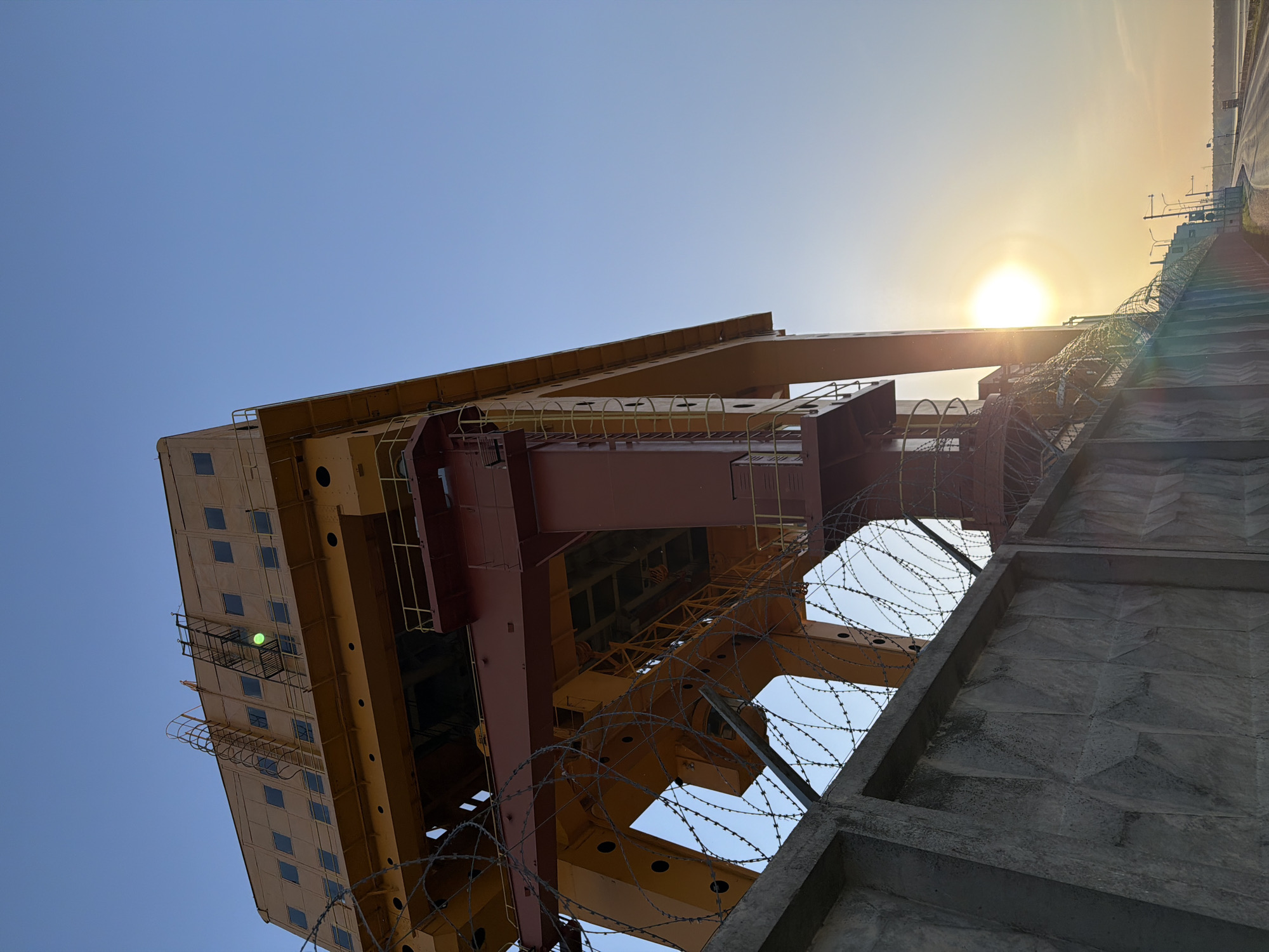 The image shows a low-angle view of a large industrial structure, likely a crane, with the sun positioned behind it, creating a lens flare effect. The structure is primarily composed of yellow and reddish-brown metal beams. A section with windows is visible on one side. Barbed wire is strung across parts of the structure and an adjacent concrete wall. The sky is a clear blue, transitioning to a lighter, sun-tinged horizon. Given the reference to Chaikovsky, Russia, it is possible this structure is located within an industrial area of that city.