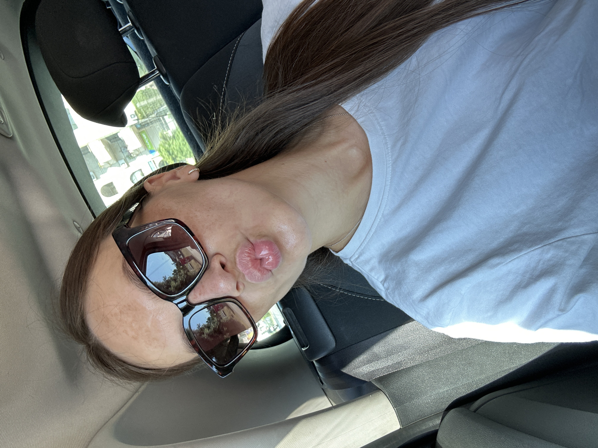 In a frontal, waist-up selfie taken inside a car, a woman with long, dark brown hair is pictured. She is wearing large, square-shaped, tortoise-shell patterned sunglasses and a plain white t-shirt. Her lips are pursed as if to give a kiss. The interior of the car is visible, including a black headrest, a seatbelt, and the back seat. Sunlight streams in from the side, casting shadows. Through the car window, buildings and trees are visible, indicating an urban or semi-urban setting. The location is identified as Donji Pobori, Montenegro.