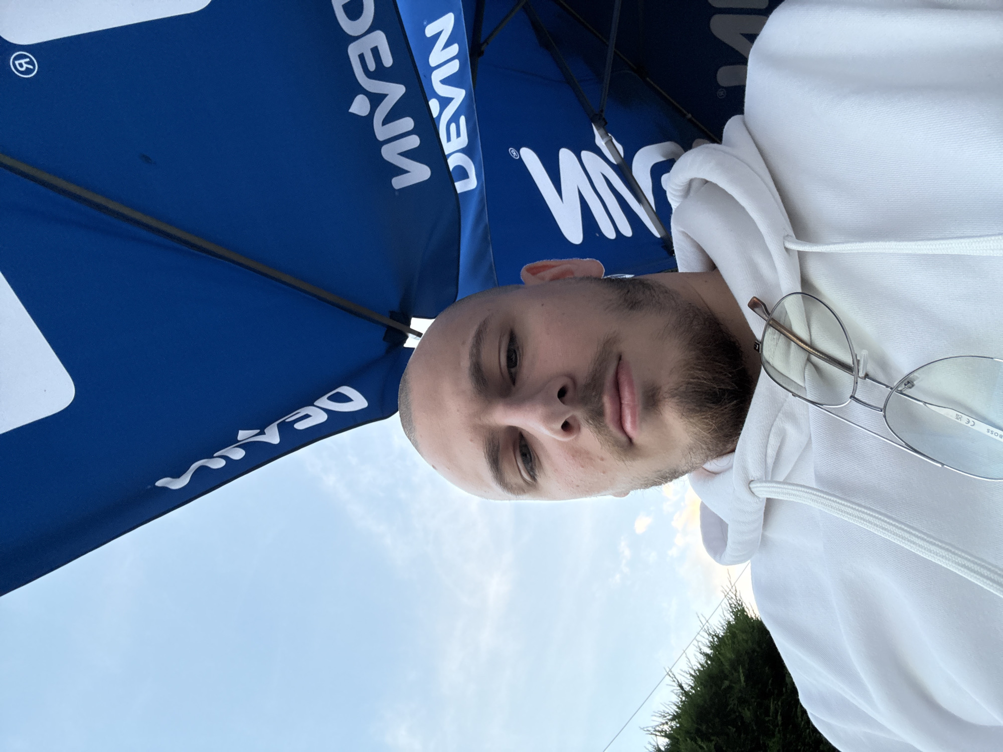 The image shows a selfie of a man wearing a white hoodie and glasses. He is standing under a blue umbrella with white lettering that reads "DEVIN." The background includes a cloudy sky and some green bushes. Without additional information, the precise location within Stara Zagora, Bulgaria, cannot be determined.