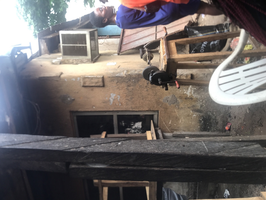 The image shows a scene in Umudike, Nigeria (latitude: 5.481, longitude: 7.546). A person wearing blue clothing and a red shirt stands on a wooden platform, looking off-camera. The platform is supporting a grinding wheel attached to a wooden table. A white plastic chair, a black bag and a piece of wood are visible on the ground, along with other debris. A window, covered by a wooden frame, is visible in the wall behind the wooden platform. The wall is made of tan concrete and has a white, rectangular object attached. The scene is partially obscured by a wooden beam in the foreground, and the top of a tree is visible at the top of the image. The image appears to have been taken from a low angle.