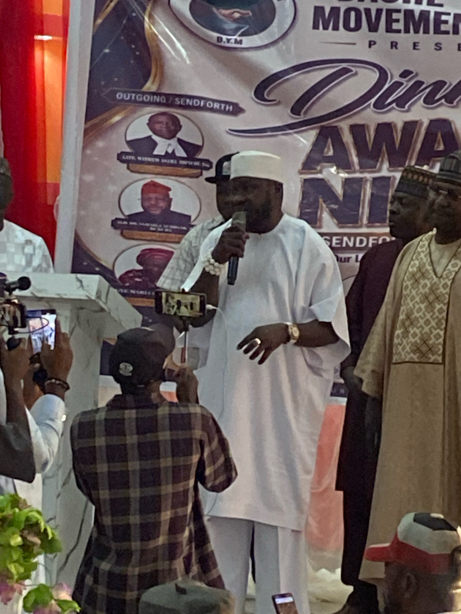 The image depicts an indoor event, likely a ceremony or gathering, with a prominent banner in the background. The banner is purple and white, featuring text that appears to be in English and possibly a local language. It announces "DINE AWARDS" and mentions "MOVEMENTS PRESENTS." There are also circular images of men on the banner, suggesting awardees or honorees.
In the foreground, several individuals are visible. A man in the center, wearing a white traditional outfit and a white cap, is speaking into a microphone and holding a gold watch on his left wrist. He is being filmed by multiple people using smartphones. To his right, another man in a light brown traditional outfit with intricate patterns on the chest is standing. To the far right, partially visible, is a man wearing a dark cap and a red and white hat.
In the lower left portion of the image, a man in a white shirt is holding a smartphone and filming. Behind him, another person is wearing a plaid shirt. Green and pink foliage is visible in the lower left corner, suggesting some decorative elements or plants at the venue. The lighting appears to be artificial, creating a somewhat warm tone. The context suggests this event is taking place in Jos, Nigeria, as indicated by the prompt.