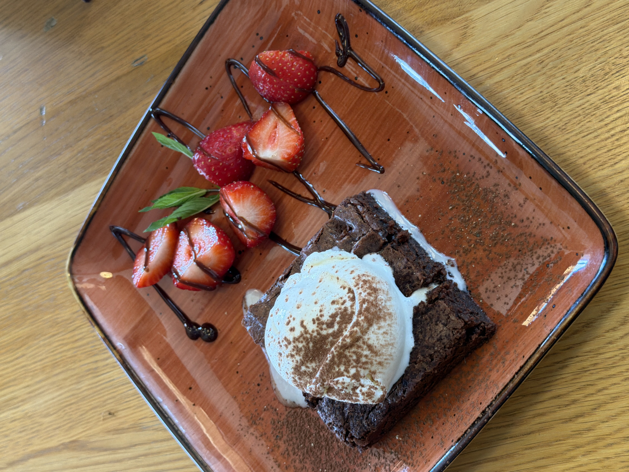 A close-up, high-angle shot shows a square, rustic brown ceramic plate on a wooden table. The plate holds a dessert, featuring a dark chocolate brownie topped with a scoop of vanilla ice cream dusted with cocoa powder. Melted white cream oozes from the side of the brownie. The dessert is garnished with several sliced strawberries and a few sprigs of mint, with chocolate sauce drizzled across the plate. The lighting is natural and bright, suggesting an indoor setting, possibly a cafe or restaurant. The wooden table is visible at the top and bottom of the frame. The scene is static, with no people or animals present. The overall impression is one of a beautifully presented dessert.