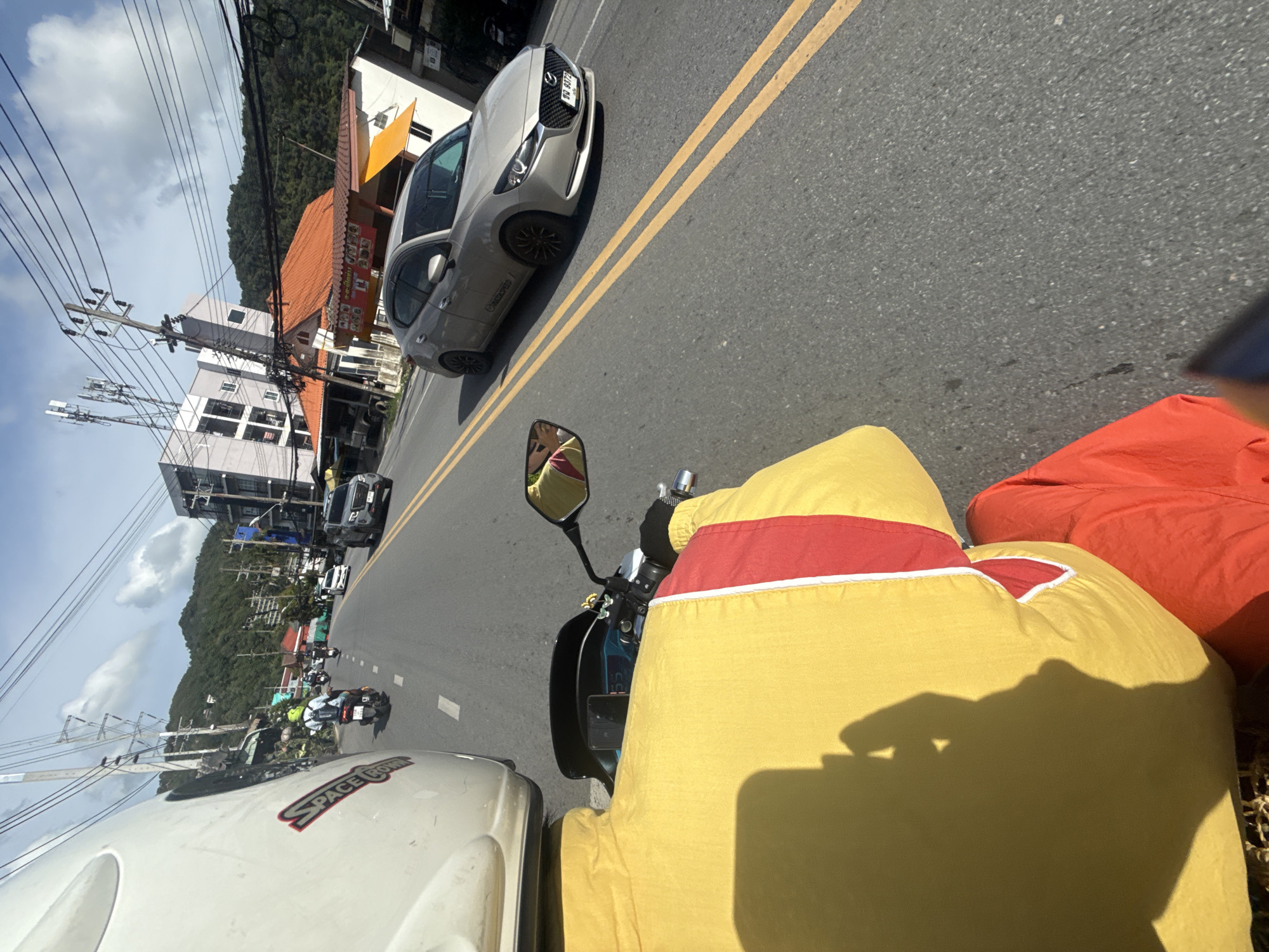 A first-person perspective shot shows a person riding a motorcycle on a road in Karon, Thailand. The rider wears a white helmet with red lettering, a yellow and red jacket, and orange pants. Ahead, several other motorcycles and a silver car are visible. The road is lined with buildings, including a hotel and shops, and lush green hills rise in the background. Power lines crisscross the sky under a bright, partly cloudy day. The car in the foreground has a license plate with Thai characters.