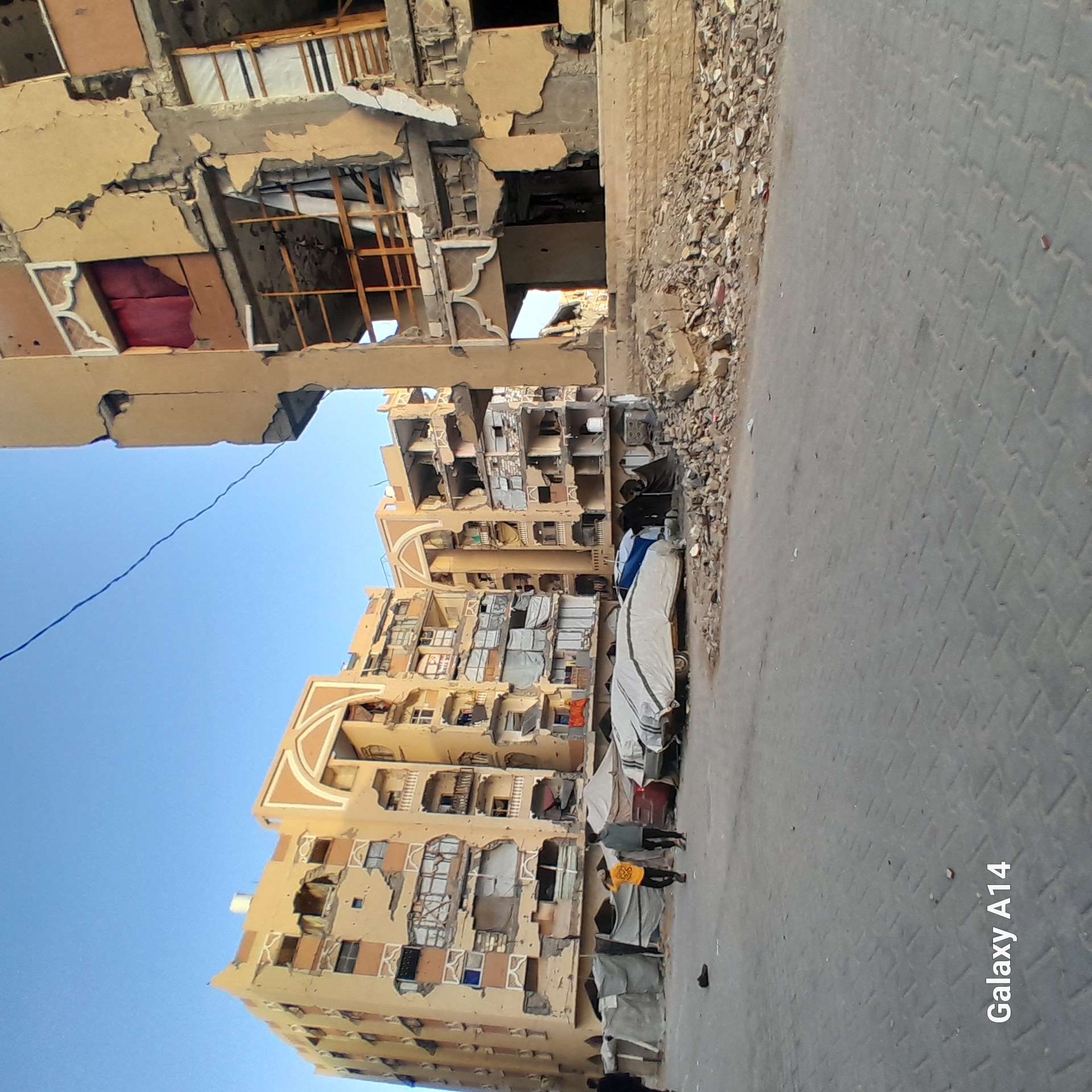 The image shows buildings in Khan Yunis, Palestinian Territory that have been severely damaged, likely from conflict or bombing. The structures have multiple floors with visible destruction, including exposed interiors, rubble, and structural damage. Debris is scattered along the roadside, and some makeshift coverings or shelters are erected against the buildings. Several people are present on the street. The sky is visible, and the overall atmosphere is one of devastation.