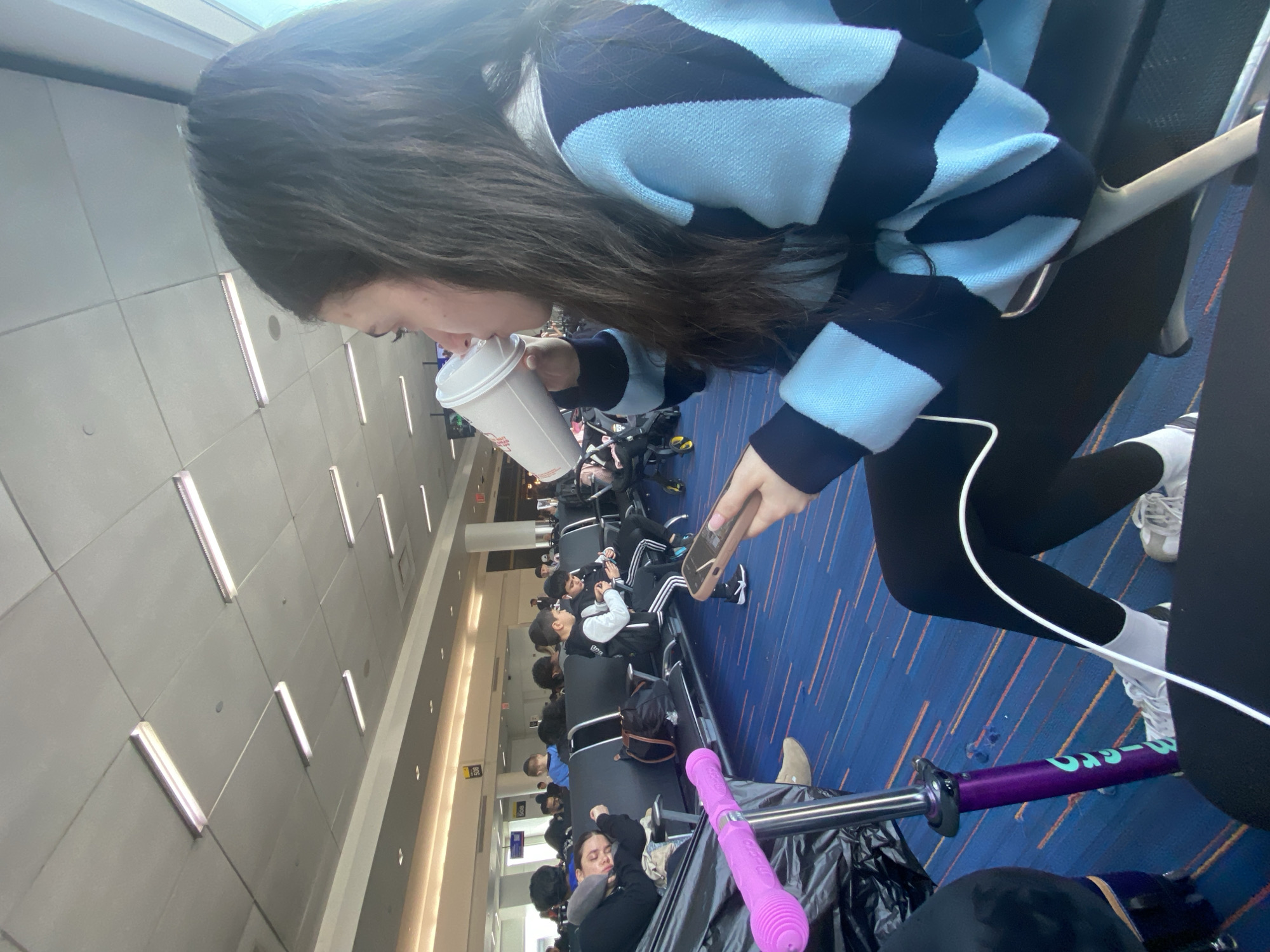 A woman with long, dark hair, wearing a light blue and dark blue striped sweater and black leggings, drinks from a white cup while holding a smartphone. She is sitting in an airport terminal, with a row of other seated people visible in the background. A purple scooter rests on the floor next to her. The location is the city of New York, United States of America.