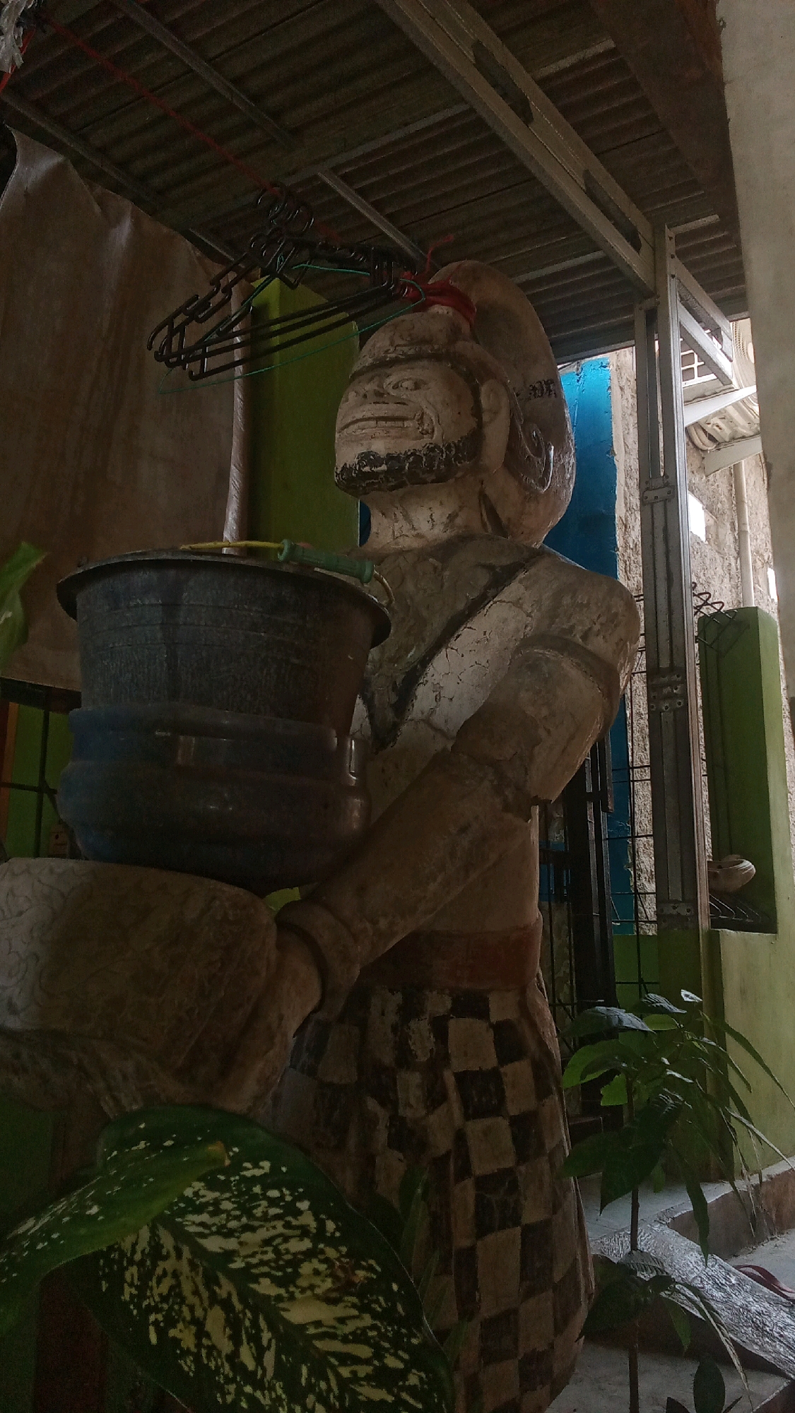 A weathered statue of a man, likely a traditional Indonesian figure, is positioned outdoors, possibly in the city of Karawang, Indonesia. The statue is made of a light-colored material, possibly concrete or stone, and exhibits signs of age and damage, with cracks visible on its surface. It is depicted holding a large, dark bucket with a green handle. The statue wears a patterned skirt, resembling a checkered design. In the background, a metal roof structure is visible, along with hanging clothes hangers. Green and blue walls frame the scene, and various plants, including large-leafed varieties, are present in the foreground and to the right of the statue. The overall setting suggests an outdoor area, potentially a courtyard or a transitional space between indoors and outdoors.