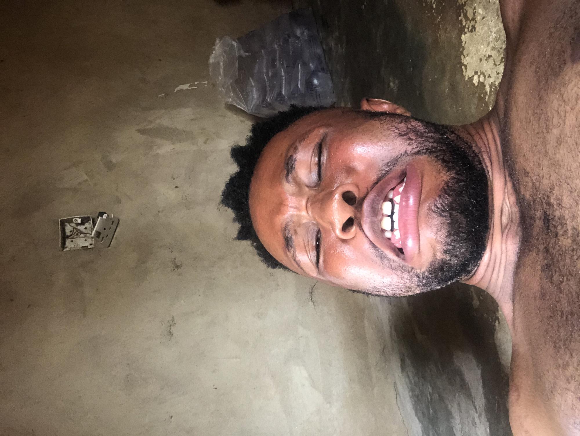 A man is lying on his side on a concrete floor. He is shirtless and has a dark complexion, a beard, and short, dark, curly hair. His eyes are closed and his mouth is open, exposing his teeth. To the left of his head is an exposed electrical outlet and a plastic bag. The setting appears to be indoors. The location is Unknown, Unknown.