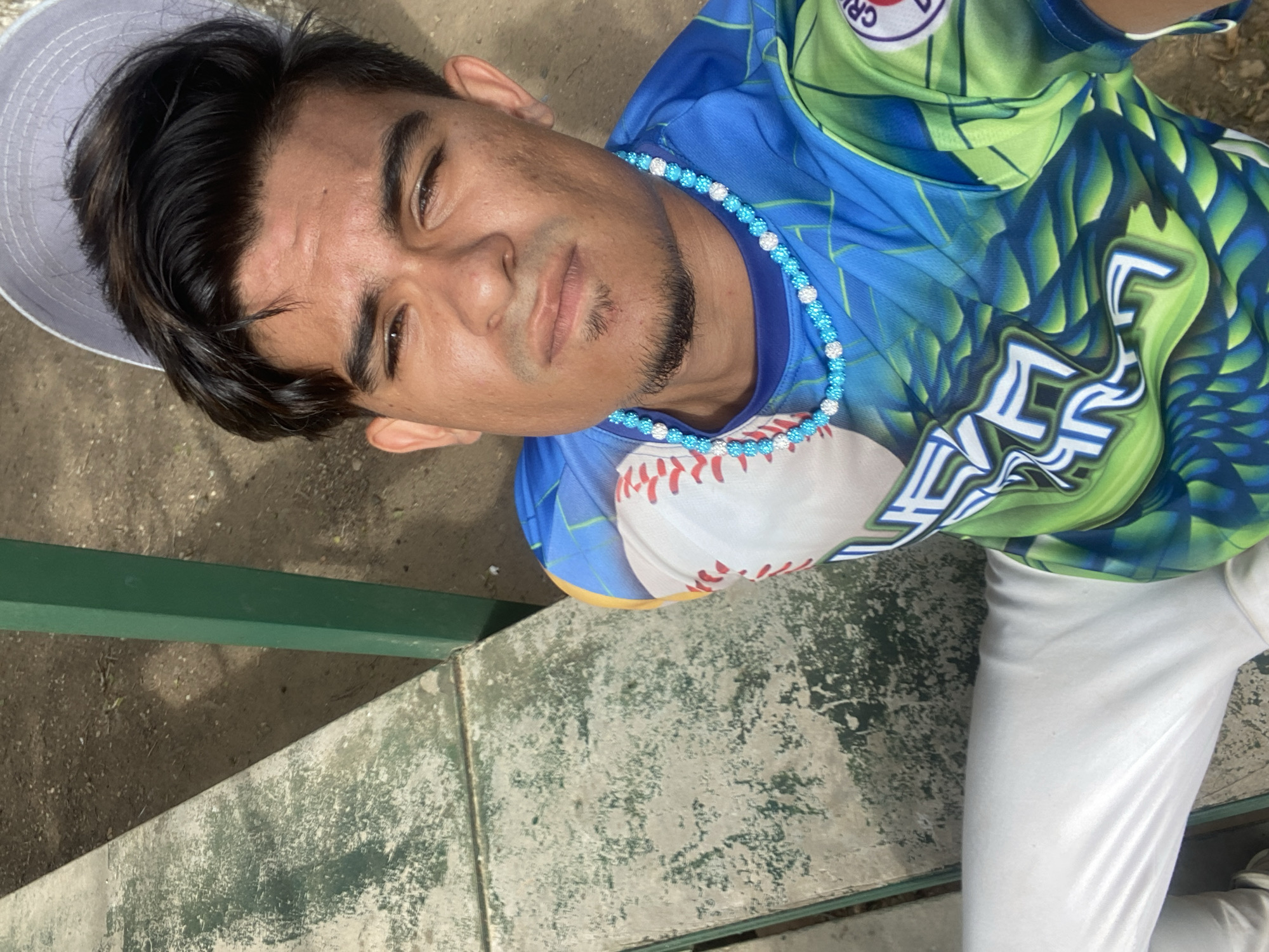A young man wearing a baseball uniform (blue, green, and white) and a light grey hat poses for a picture. He wears a blue beaded necklace with white accent beads. He is seated on a bench made of concrete and green painted metal in what appears to be an outdoor location. The concrete has a weathered appearance. The location is Las Piedras, Venezuela.