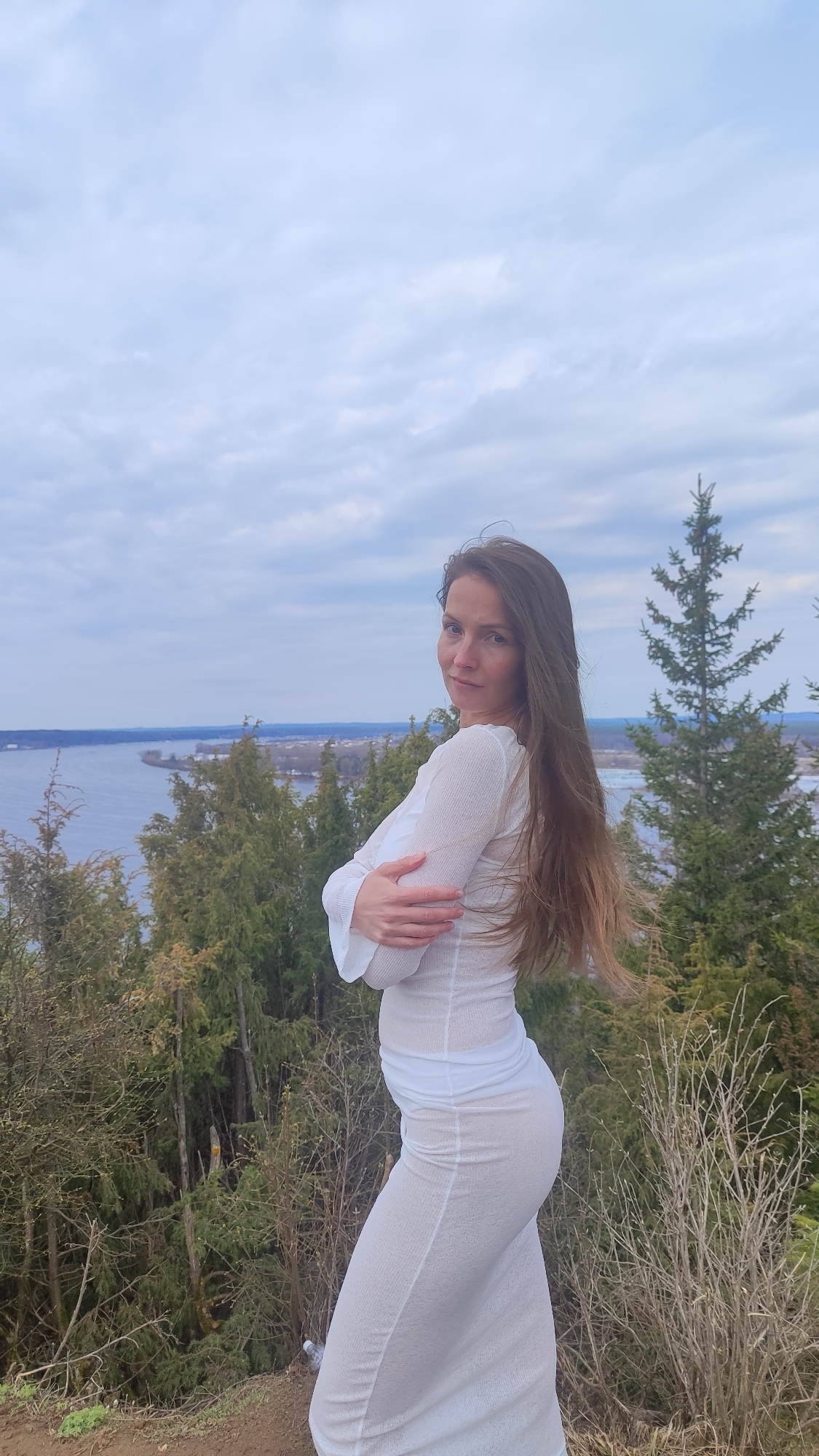 A woman stands on a hill in Ольховка, Russia. She has long brown hair and is wearing a long, white, form-fitting dress. She is facing the camera with her arms crossed. Behind her is a landscape with trees, a body of water, and a cloudy sky.