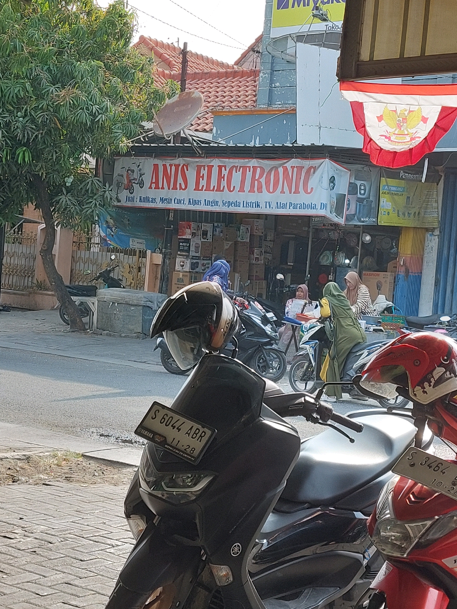 The image captures a street scene in Sumuragung, Indonesia, dominated by the storefront of "ANIS ELECTRONIC." The shop displays a banner with the business name in large red lettering, accompanied by illustrations of a scooter and a fan. Below the name, text in Indonesian lists items for sale: "Jual: Kulkas, Mesin Cuci, Kipas Angin, Sepeda Listrik, TV, Alat Parabola, Di." This translates to "For Sale: Refrigerator, Washing Machine, Fan, Electric Bicycle, TV, Satellite Dish, Etc."
Several motorcycles are parked in front of the shop. A dark-colored scooter with a license plate reading "S 6044 ABR" is prominent in the foreground. Another motorcycle, partially visible, has a red front fairing and a license plate that reads "S 3464 B". On the right side of the image, a red motorcycle with a distinctive decal is also visible.
Three women are present near the shop entrance, two of whom are wearing headscarves. One woman in a green hijab is seated on a scooter, while another woman in a pink patterned outfit is seated on a stool. A third woman, also wearing a headscarf, is standing behind a counter within the shop.
The environment is a street with a paved sidewalk. A large tree with green foliage stands to the left of the shop. A satellite dish is mounted on the roof of the building. An Indonesian flag, folded and displayed, is also visible above the shop entrance. The buildings have red tiled roofs and some display signage, including "Panasonic" and a yellow sign with Indonesian text and graphics. The overall impression is of a bustling local business in a street setting.