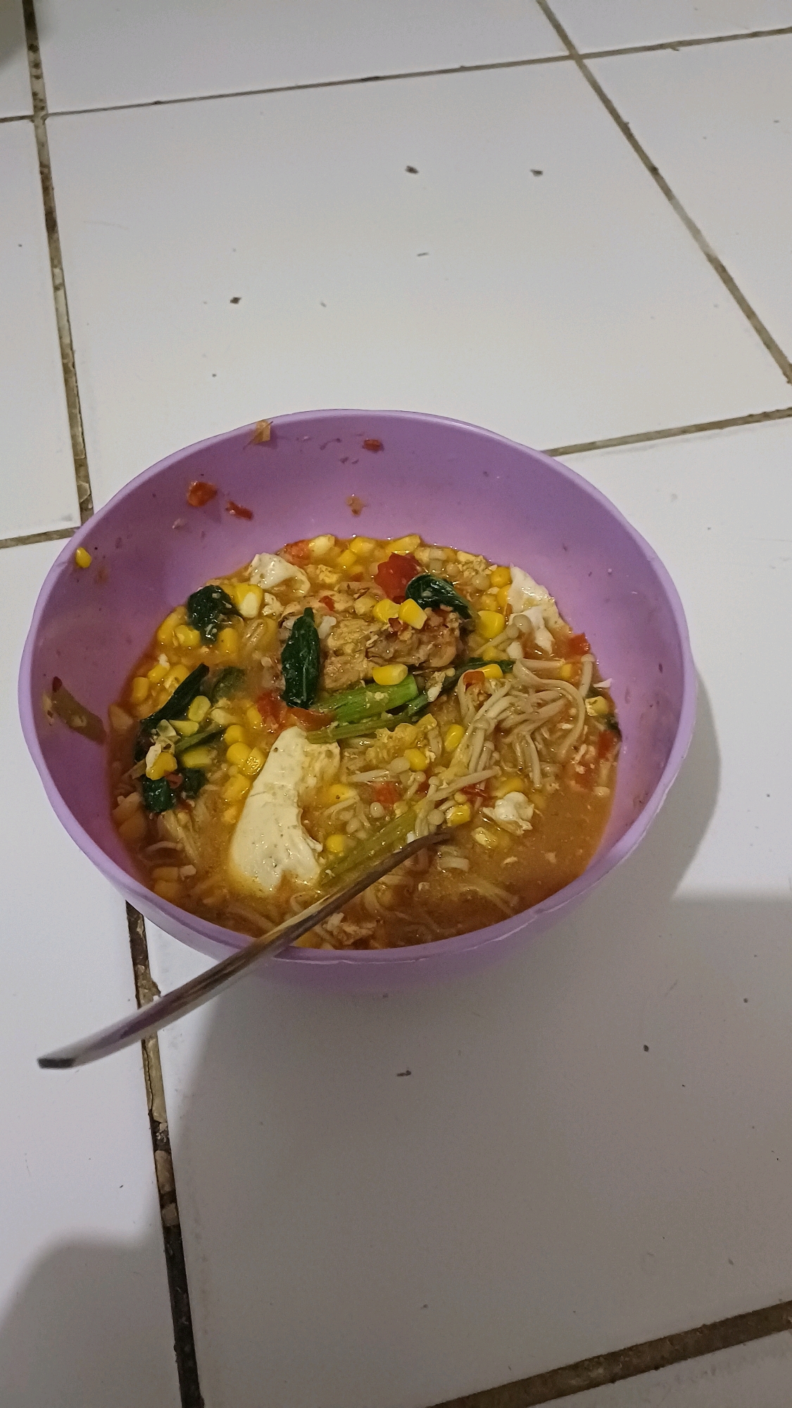 A close-up, high-angle shot shows a purple bowl filled with a noodle soup dish. The dish contains enoki mushrooms, corn, chopped tomatoes, green leafy vegetables, and pieces of chicken and tofu. A metal spoon is resting inside the bowl, partially submerged in the soup. The bowl is placed on a light-colored tiled surface, with dark grout lines visible between the tiles. The lighting suggests an indoor setting. The location is identified as Gunung Putri, Indonesia.