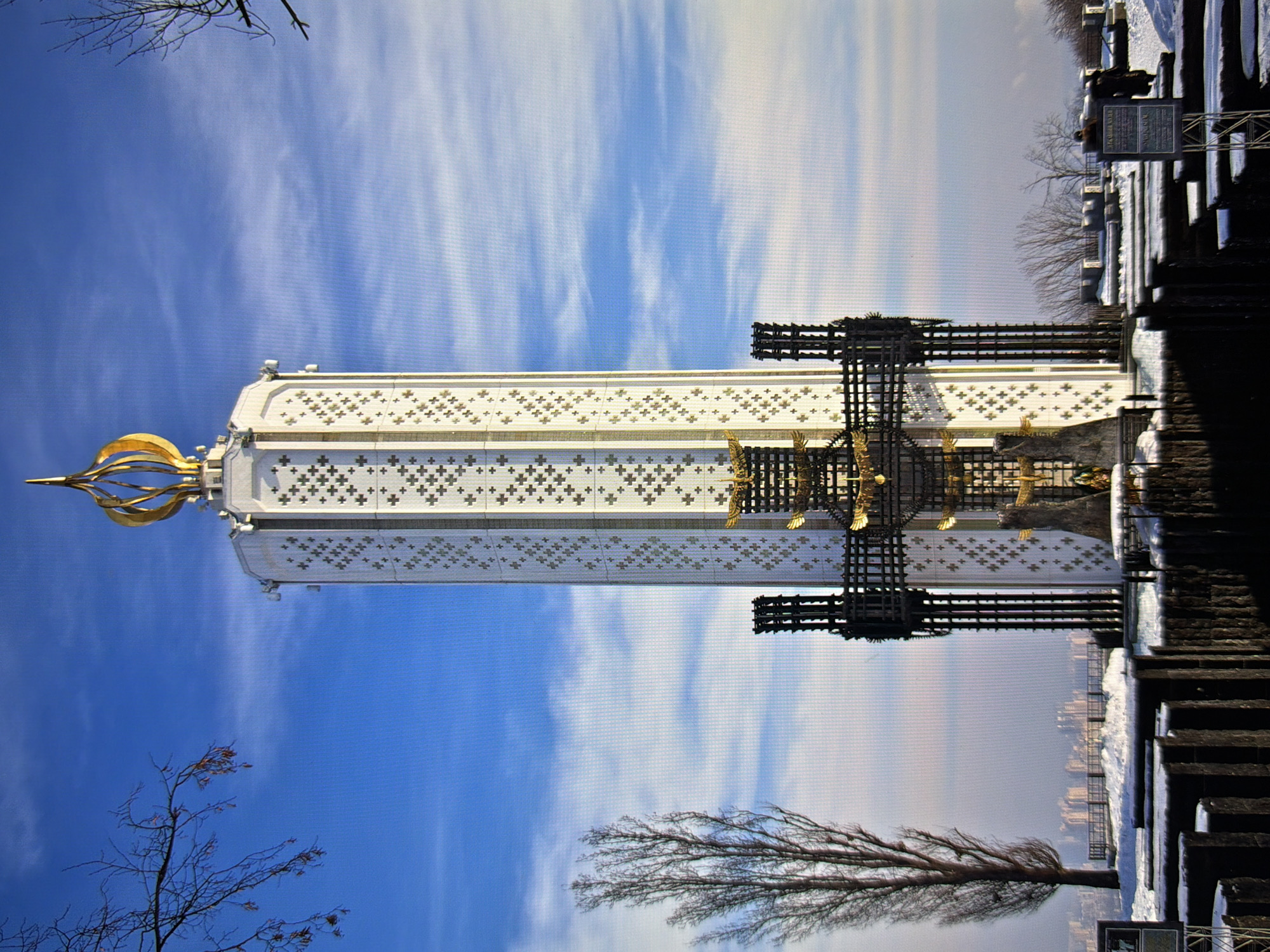 The image depicts the Memorial of the Holodomor in Kyiv, Ukraine. It is a tall, white, candle-shaped monument adorned with decorative crosses and topped with a golden flame. At the base of the monument is a large, black, cross-shaped structure with golden eagle motifs. The memorial is situated in a snowy landscape with bare trees and a distant cityscape visible. The sky is blue with scattered clouds. In the foreground, there are dark, stone-like structures, possibly part of the memorial complex, also covered in snow. Two individuals are visible in the middle ground, standing near informational signs. The overall scene is solemn and reflective, set against a bright, clear day.