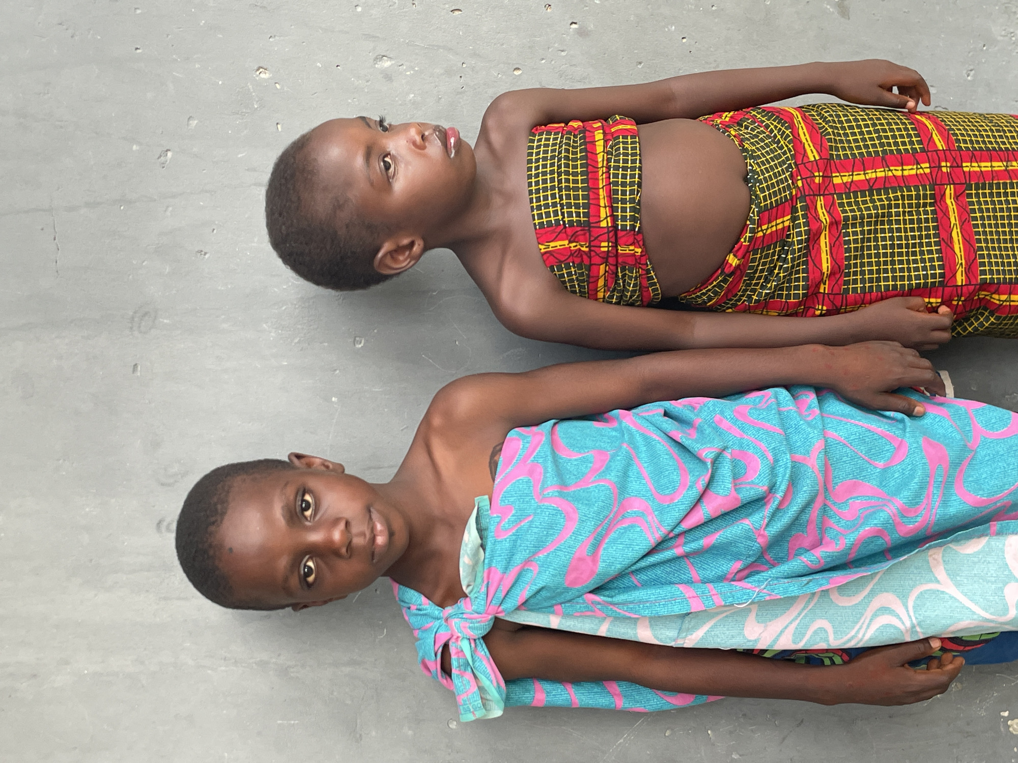 Two young children are depicted lying on a light grey, textured concrete or paved surface. The image is taken from a slightly elevated perspective, with the children oriented diagonally across the frame. The lighting is bright and even, suggesting daytime.
The child positioned further from the camera is partially dressed in a vibrant red, yellow, and black patterned fabric, possibly a traditional African print, wrapped around their chest and lower body. This child has short dark hair and is looking upwards and to the right with a neutral to contemplative expression.
The child closer to the camera is wrapped in a different fabric, predominantly turquoise-blue with bright pink swirling patterns, tied around their left shoulder. This child also has short dark hair and is looking directly at the camera with a calm, neutral expression. Both children have dark skin tones.
The scene is simple, with no other objects or background elements visible beyond the ground surface. The children appear to be resting or posing. No specific activity is taking place. There is no visible text in the image. The clothing patterns suggest a cultural context typical of the region, aligning with the provided location of Badagry, Nigeria.