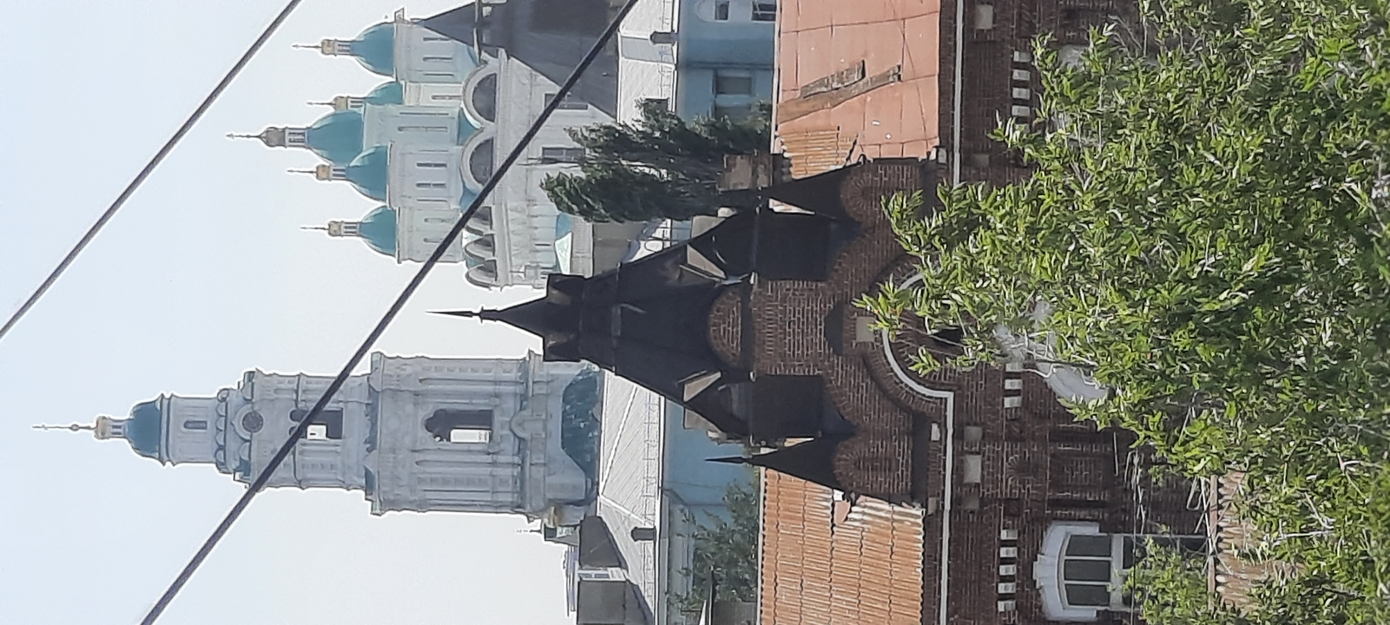 The image shows a view of the Astrakhan Kremlin complex in Astrakhan, Russia, with a prominent bell tower and several domed cathedrals. In the foreground, a brick building with a dark, pointed roof and rusty corrugated iron roofing is visible. Wires are present across the top of the image. Green trees partially obscure the view. The buildings are illuminated by daylight under a clear sky.