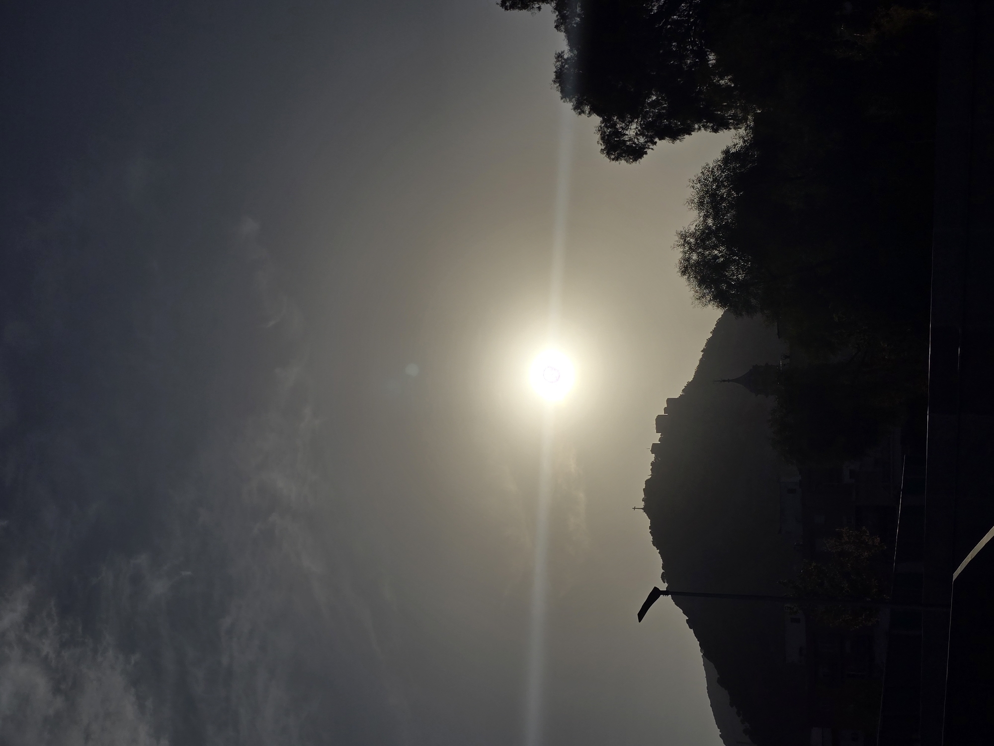 The image shows a silhouetted view of the Santa Catalina Castle on a hill overlooking Jaén, Spain. The sun is setting or rising, creating a bright lens flare across the image. A cross is visible on the top of the hill. In the foreground, there are dark silhouettes of trees and buildings. A street lamp is also visible on the left side of the image. The sky is filled with hazy clouds, and the overall mood of the image is atmospheric and slightly dramatic.
