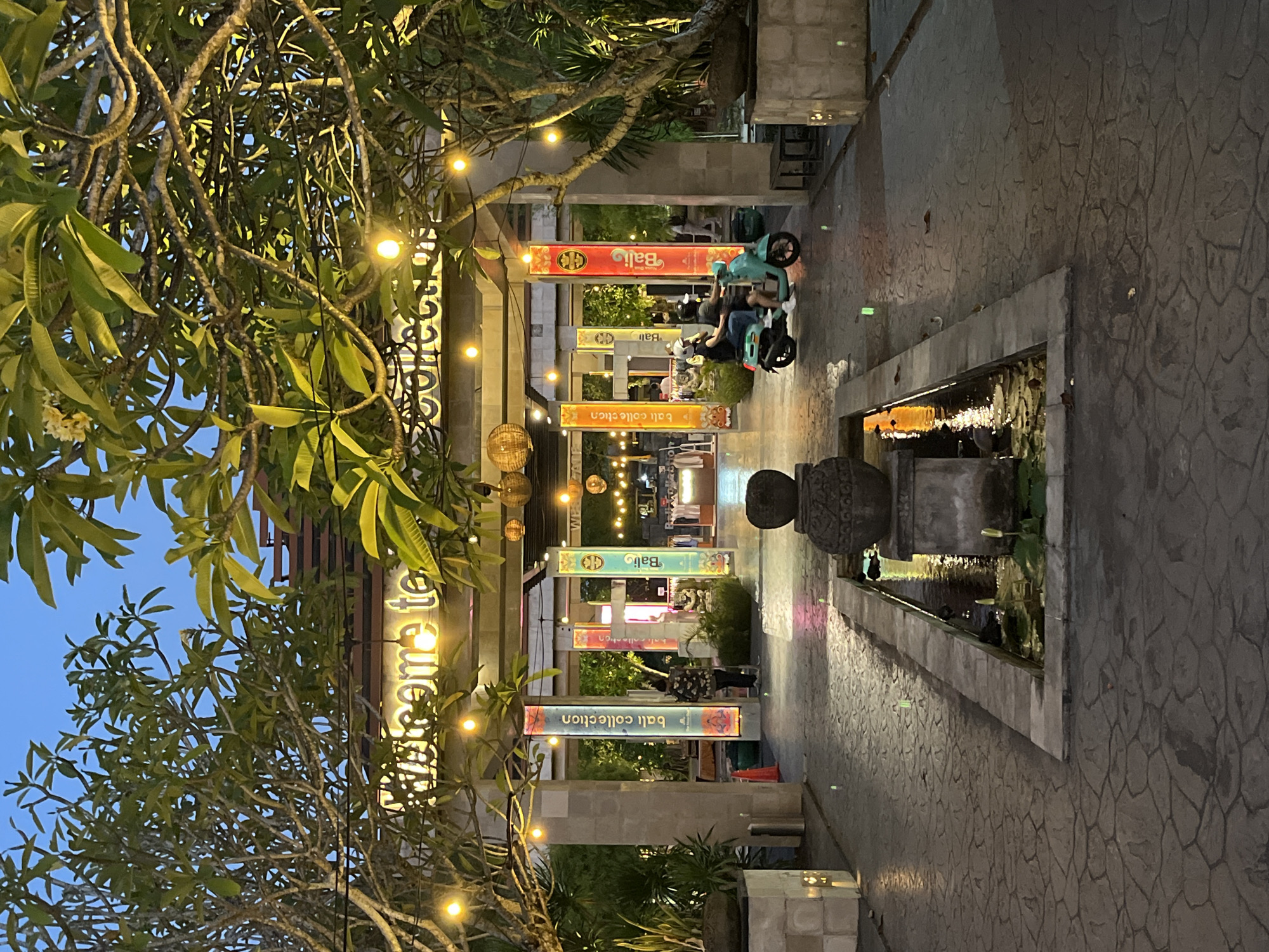 The image shows an outdoor scene in Bali, Indonesia, likely at dusk or early evening. A stone pathway extends into the distance, bordered by tropical plants and architectural elements. Overhanging the path are structures with banners displaying "Bali Collection" in multiple languages. Strings of lights illuminate the area, and people are visible in the background, some riding electric scooters. A stone fountain or water feature sits on the right side of the pathway. The sky is visible in the upper-left corner, hinting at the fading daylight.
