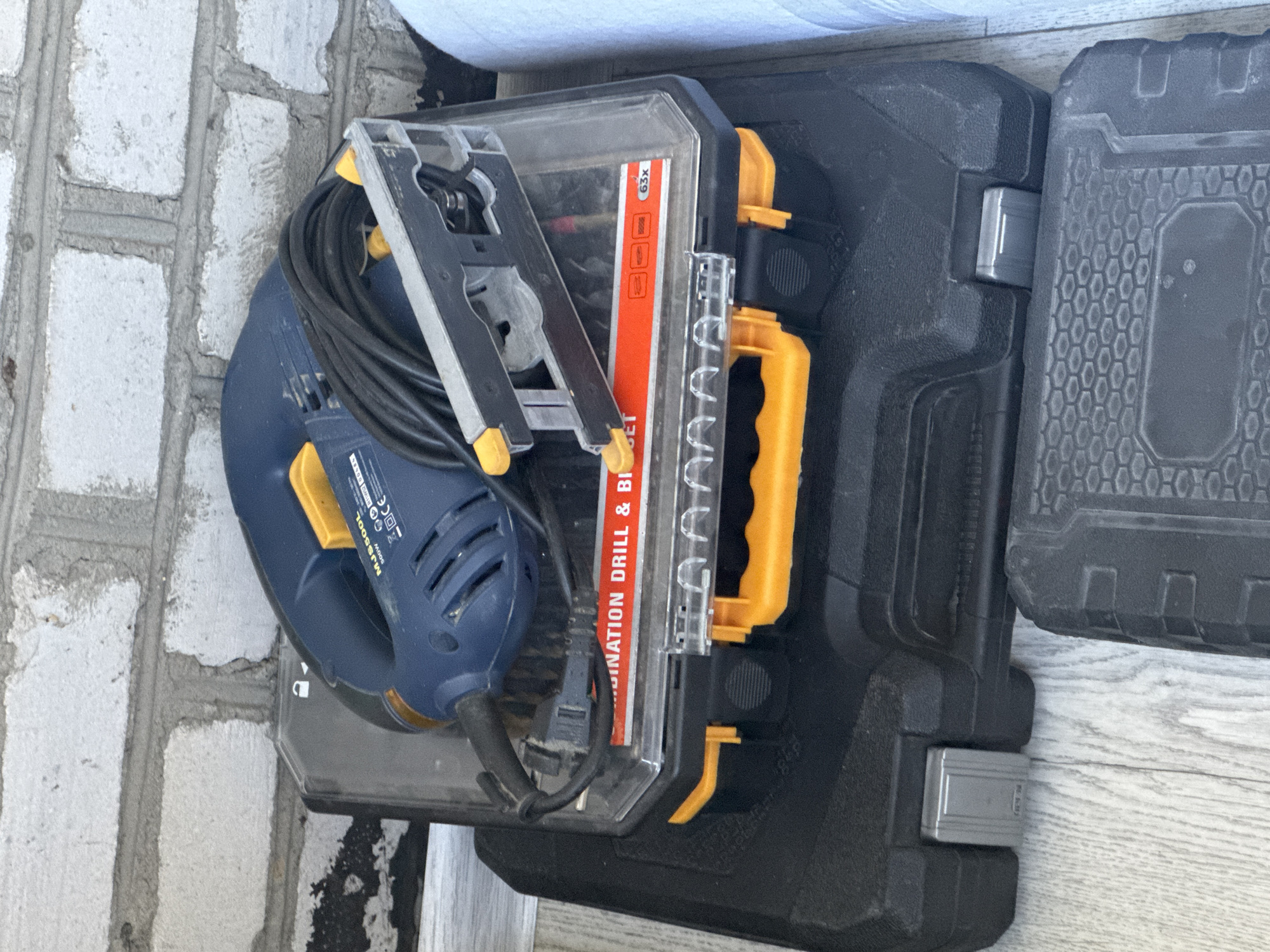 This image shows a close-up, overhead view of a jigsaw and its black plastic carrying case, placed on a light-colored wooden floor. The jigsaw is blue and yellow, with a black power cord coiled on top. The carrying case is black and has a distinctive textured pattern on its lid, with a yellow handle. Part of another similar black case is visible to the right.
Visible text on the jigsaw reads "MJS 500L 500W". There is also a red and orange label on the jigsaw's plastic guard with text that includes "INATION DRILL & BI" and "63x".
The setting appears to be indoors, possibly a workshop or garage, given the presence of tools and the floor material. There are no people visible in the image. The lighting suggests it could be daytime, though the time of day and weather are not discernible from the image itself. The overall scene is a still life of power tools. The location context provided is "Яромінскі сельскі Савет, Belarus".