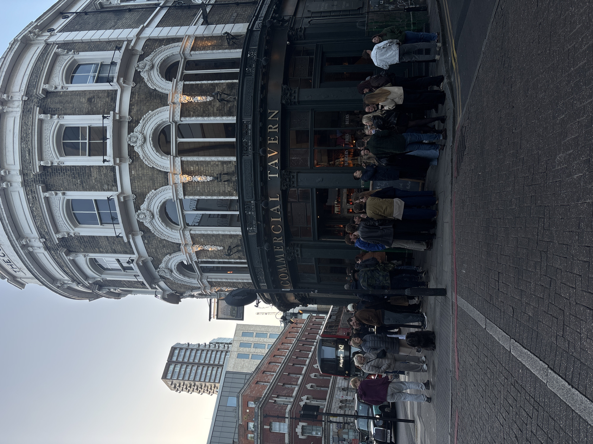 A street view captures "The Commercial Tavern" in London, United Kingdom. People are queuing outside. A bus marked "Aldgate 242" is visible behind the queue. A black cab is in front of them. Nearby buildings of varying heights and architectural styles form the backdrop.