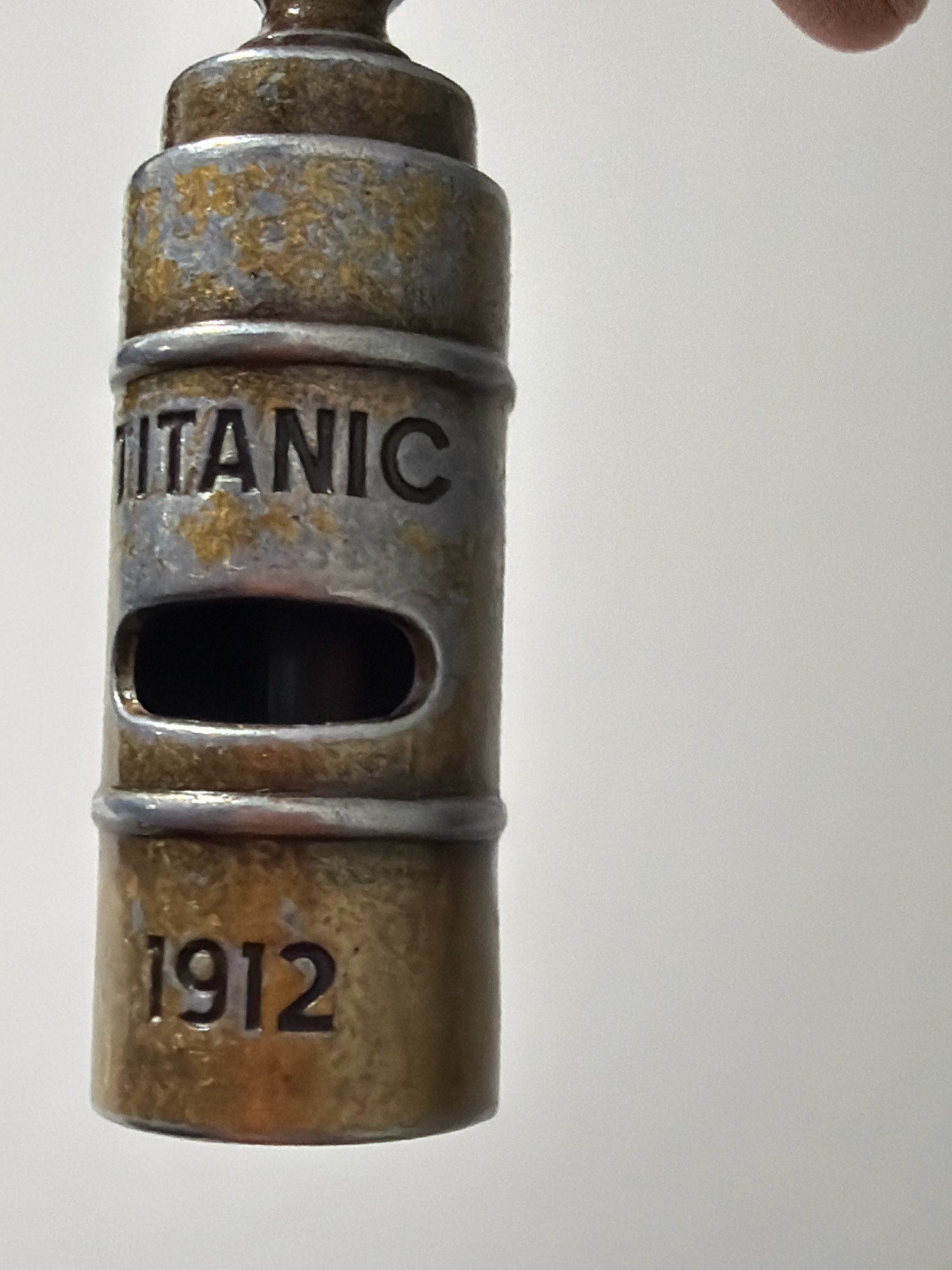 The image shows a close-up, eye-level, vertical shot of a brass whistle with the words "TITANIC" and "1912" inscribed on it. The whistle has an oval opening in the center. There is some wear and tear visible on the surface of the whistle, with patches of discoloration and patina. A hand is partially visible at the top right corner of the image, suggesting someone is holding the whistle. The background is a plain, light gray surface. The image was taken indoors with neutral lighting against a solid backdrop. There is no discernible connection to Wrocław, Poland.