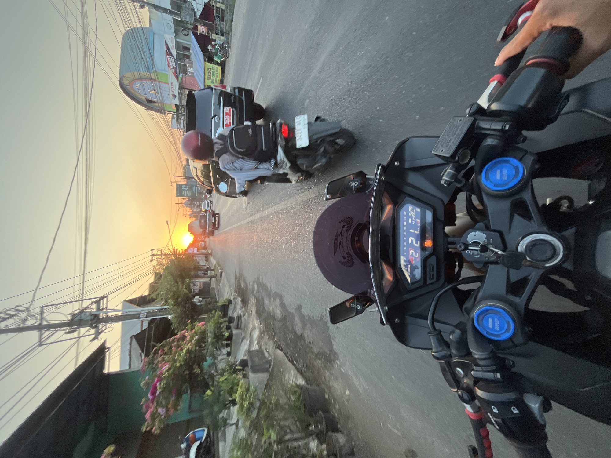The image depicts a first-person perspective from a motorcycle traveling on a street in Margoyoso, Indonesia, during sunset. The road is paved and appears to be a main thoroughfare with traffic. On the left side of the road, there are buildings with green facades and potted plants with flowering bushes. Overhead, numerous electrical wires crisscross the sky, typical of Indonesian streetscapes.
In the foreground, the handlebars and digital dashboard of the motorcycle are visible, showing a speed of 21 kph. The dashboard also displays what appears to be time and gear indicators.
On the road ahead, a motorcycle is visible with a rider wearing a red helmet and a backpack. This motorcycle is in front of a black SUV. Further down the road, other vehicles and a truck are present, moving in the same direction. The setting sun casts a warm glow, illuminating the sky and reflecting on the wet asphalt. The overall scene conveys a sense of daily life and commuting in a town or city in Indonesia.