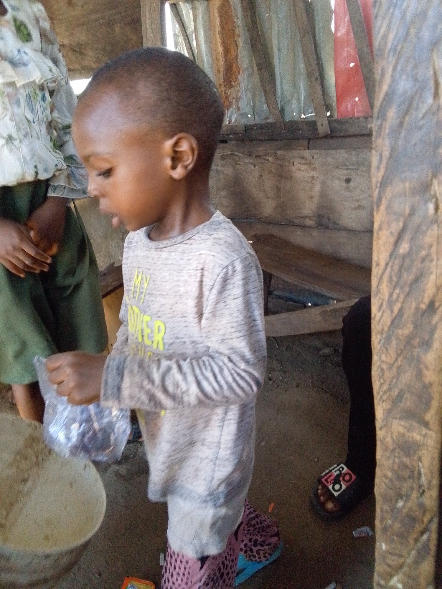 A young child, likely a boy, is the central subject of the image, depicted from the waist up in a medium shot. He has short, dark hair and is wearing a light gray long-sleeved shirt with some light yellow text, partially visible as "MY OTHER". He is also wearing distinct pink and blue patterned slip-on shoes or sandals. The child is holding a small, clear plastic bag with his right hand, seemingly looking down at its contents or at a light-colored, possibly ceramic, bowl filled with a light-brown powder or flour, which is visible in the lower left foreground. His mouth is slightly open, suggesting he might be speaking or focused intently.
Behind and to the left of the child, another person is partially visible, wearing a white ruffled top with a floral pattern and a dark green skirt or trousers. Their hands are clasped or holding a small, orange-colored object.
The setting appears to be an indoor or semi-enclosed space with a dirt floor. The background consists of rough wooden planks and support beams, along with corrugated metal sheets, some of which show signs of rust and wear, creating a rustic or makeshift environment. A glimpse of red material is seen behind the metal sheets. In the lower right, the foot of another person is visible, wearing a black sandal with a white and red pattern that includes the letters "LO" and possibly "8O". The lighting suggests it is daytime, with natural light filtering into the somewhat dim space. The overall atmosphere is mundane, capturing an everyday moment with no strong emotions expressed.