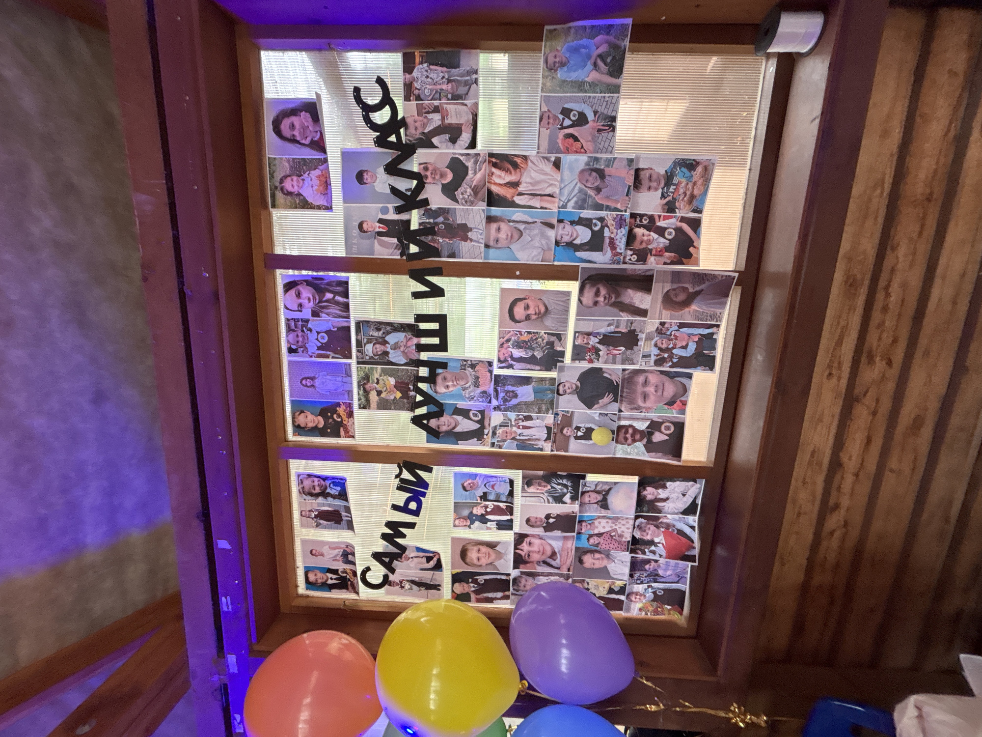 The image shows a window frame with a display of student photos, arranged in rows and columns. The Cyrillic text "САМЫЙ ЛУЧШИЙ КЛАСС" (meaning "The Best Class") is spelled out in cut-out letters and arranged vertically on the left side of the window. Below the window, there are several inflated balloons in orange, yellow, purple, and blue. The frame and the surrounding walls appear to be made of wood. A partially visible ceiling or roof structure is present. The overall environment seems to be an indoor setting, possibly a school event or party. A location of Chaikovsky, Russia is specified.