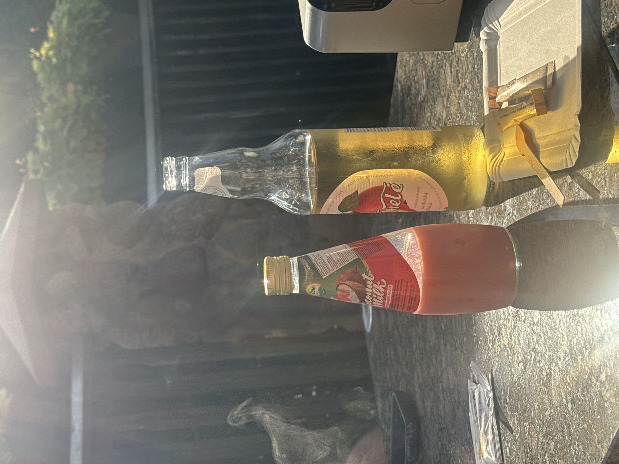 In Rūdiškės, Lithuania, on a sunlit day, a table outdoors displays two beverages and a paper tray. To the left is a conical glass bottle containing a pink, opaque liquid, labeled "Coconut Milk Strawberry Flavor". Next to it stands a clear glass bottle filled with a yellow, translucent liquid, with a label featuring a red apple and the word "Nirle" (likely misspelled and should be "Nirvelė" or similar in Lithuanian). The bottle appears to be a type of apple cider or juice. On a disposable paper tray to the right, two small packets, possibly sugar or sweetener, and a small wooden stirrer are arranged. The table surface is textured, resembling granite. In the background, out-of-focus foliage and a dark, textured wall are visible. The overall impression is a casual outdoor refreshment setting.