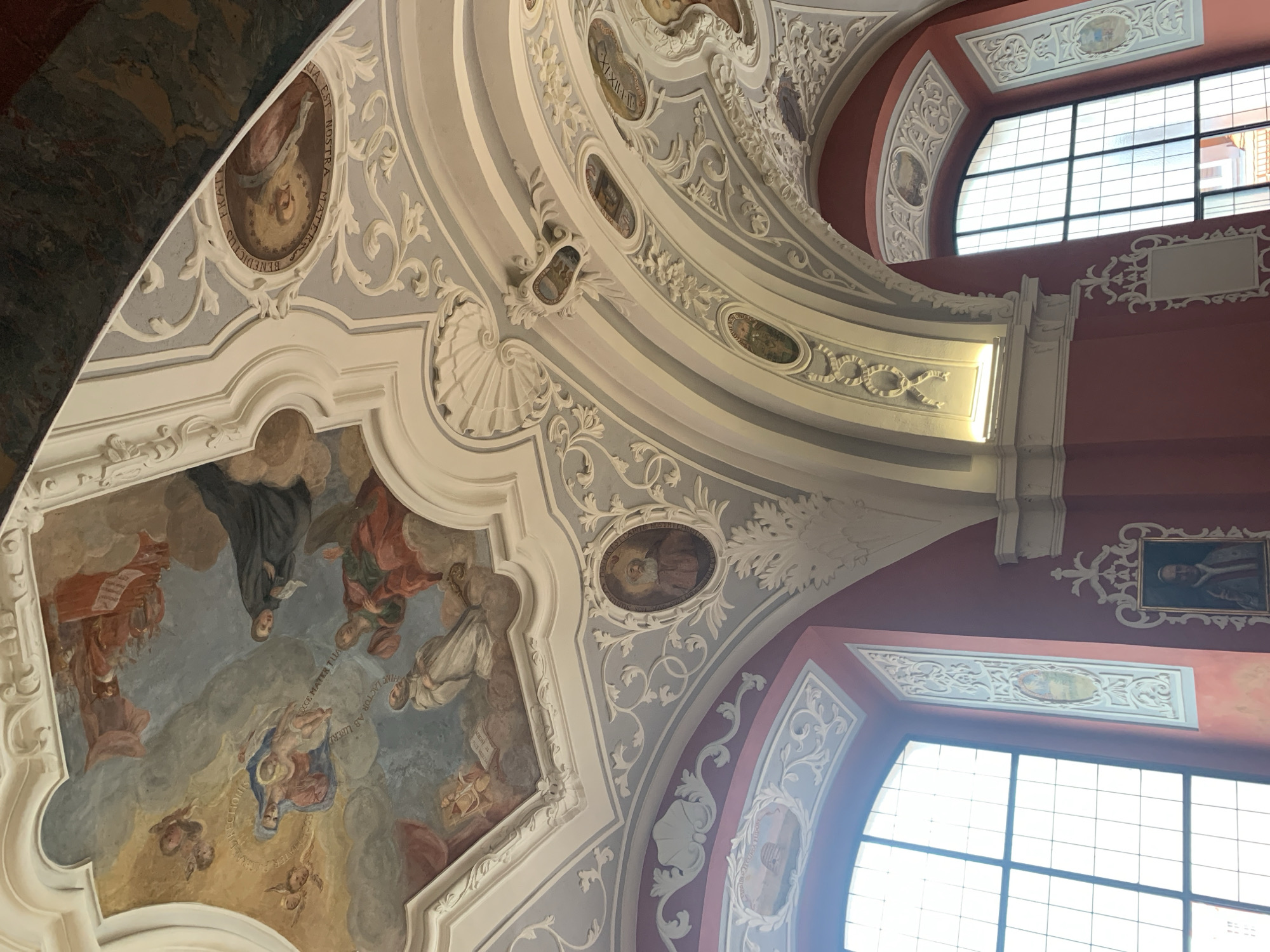 This image depicts the interior of the historic Municipal Library in Poznan, Poland, specifically the ceiling and upper walls of one of its halls. The ornate Baroque ceiling is adorned with elaborate frescoes and stucco decorations. The frescoes depict religious scenes with numerous figures, including cherubs, angels, and what appear to be saints, rendered in a soft, classical style with a muted color palette.
The ceiling is characterized by its vaulted structure and is divided into various panels and sections, each featuring painted narratives or decorative motifs. White stucco work, with intricate scrollwork, shells, and floral elements, frames the frescoes and adds depth and texture to the ceiling. Insets within the stucco contain smaller oval paintings, some of which appear to be portraits.
Below the ceiling, two large, arched windows with grid-like panes are visible. The walls beneath the windows are painted a rich reddish-pink color. On these walls, there are framed paintings and decorative stucco elements that echo the motifs found on the ceiling. One prominent painting on the left wall shows a figure in religious attire. The overall impression is one of grandeur and artistic opulence, typical of Baroque ecclesiastical or public architecture. The lighting appears to be a combination of natural light from the windows and artificial lighting, casting gentle illumination on the decorative surfaces.