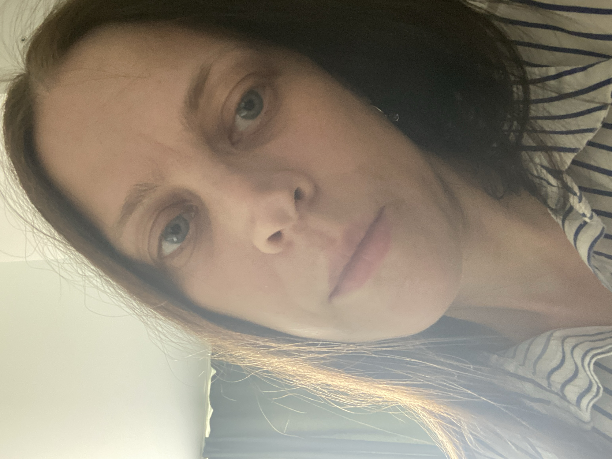 A close-up, low-angle selfie of a person with medium-brown hair and fair skin is captured. The individual is wearing a white and blue striped collared shirt. Their left eye is clearly visible, and their mouth is slightly parted, with a faint smile. The lighting suggests an indoor setting, with a window or light source visible on the left side of the frame, casting a warm glow on the person's hair and face. A portion of a light-colored wall and a dark curtain are discernible in the background. The image is a direct, unposed photograph of the person's upper body and face, taken from a close perspective. The location is not specified in the image.