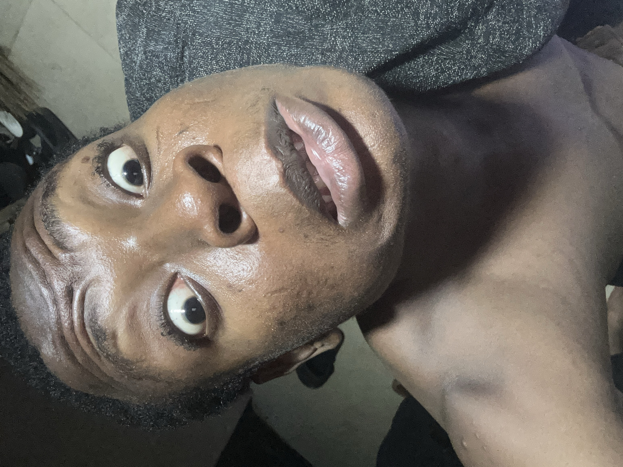 A medium close-up shot depicts an individual with dark skin, positioned at an unusual angle, appearing as if they are lying down with their head tilted towards the camera. Their eyes are wide, with a slight sheen, and their lips are parted. The skin has a glossy appearance, suggesting moisture or sweat. Some stubble is visible. The individual is partially clothed with dark grey fabric near the top of the frame. The background is out of focus, contributing to the emphasis on the individual's face.