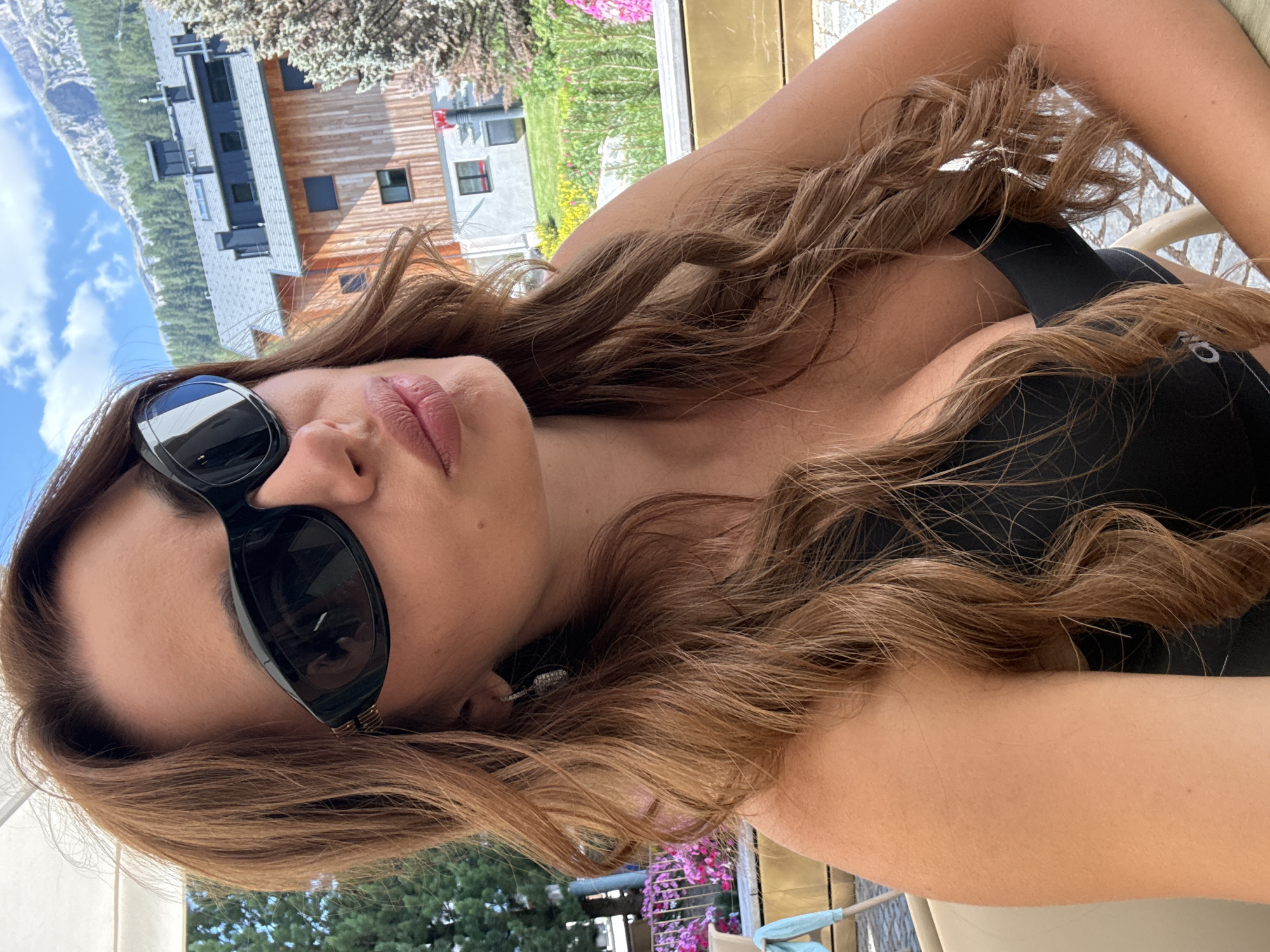 In Calfosch - Kolfuschg - Colfosco, Italy, a woman with long, wavy, light brown hair is pictured from the chest up, taking a selfie. She is wearing black sunglasses and a black top. Her lips are pursed and her head is tilted slightly to the right. Behind her, a building with a wooden exterior and modern architecture is visible, with trees and mountains in the background under a blue sky with white clouds. The setting appears to be an outdoor seating area, with hints of pink flowers and a yellow railing.