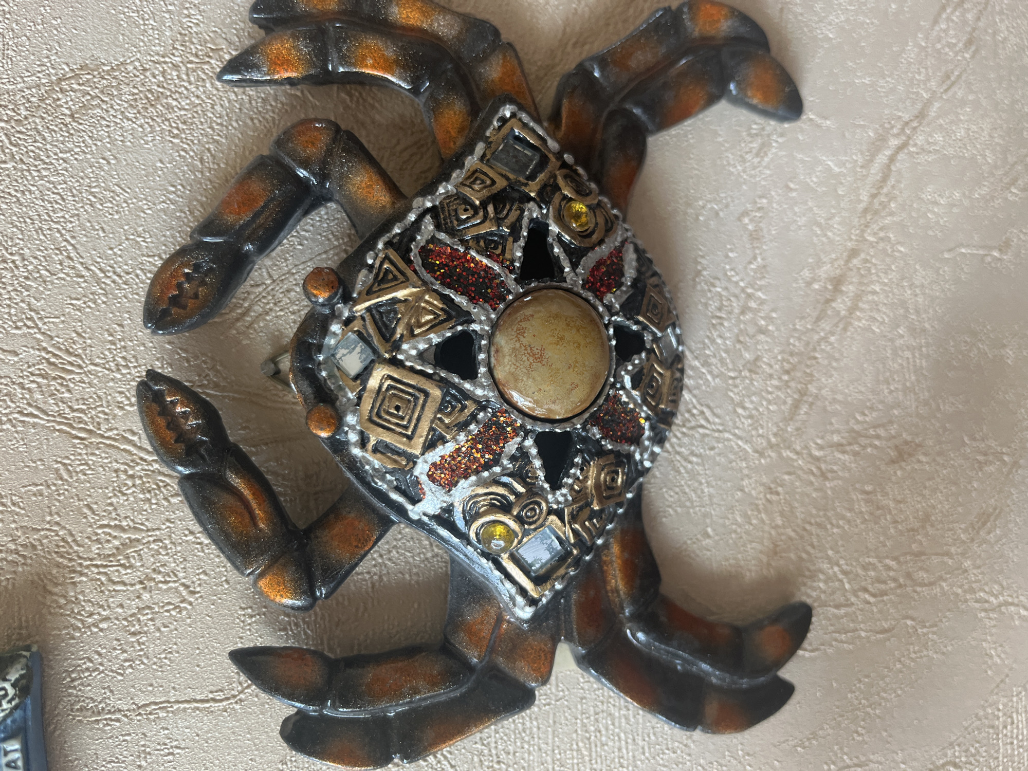 The image shows a decorative crab figurine on a textured, light-colored wall. The crab is dark brown and orange with intricate details on its shell, including gold geometric patterns, red glitter, and small gemstones. The background wall appears to be a typical interior wall with a textured finish. There is no indication of the city of Vladimirovka, Russia, or any specific location within a city. The scene is static and does not depict any actions or interactions.