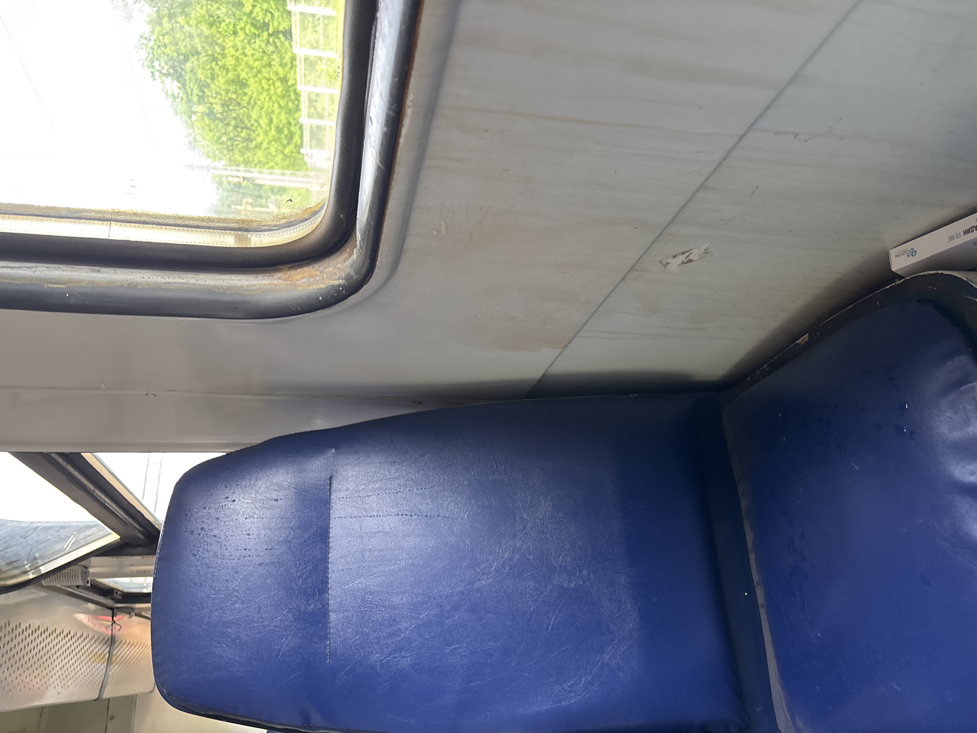The image depicts the interior of a passenger train, possibly in Moscow, Russia. A blue vinyl seat is in the foreground, with a window and the train's interior walls visible in the background. The window frame appears weathered. The environment suggests public transportation within a city.
