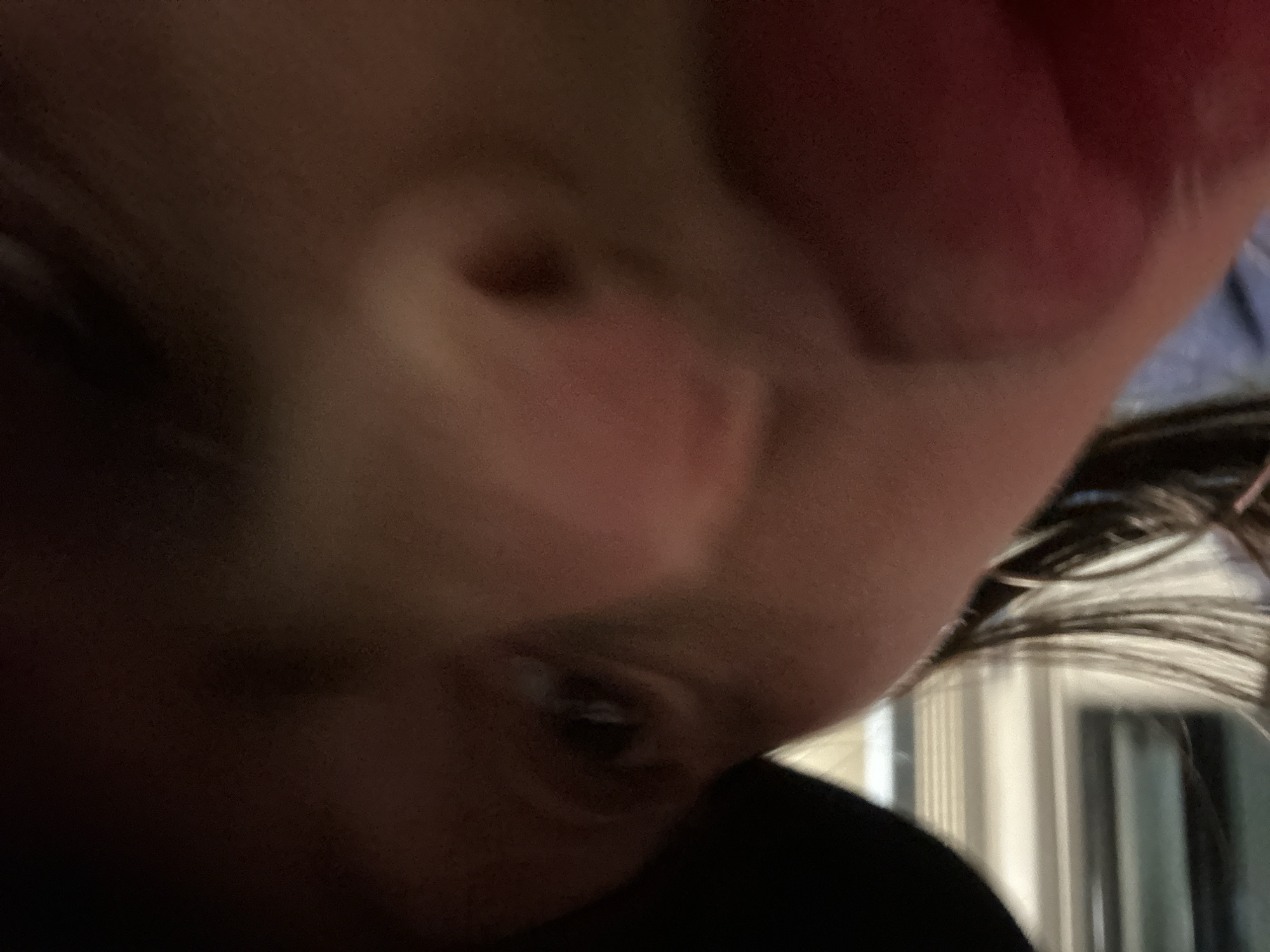 This image depicts a close-up, low-light, slightly out-of-focus portrait of an individual's face, taken from a low angle. The subject's left eye is clearly visible, dark, and appears to be looking directly at the camera. The central part of the face, including the bridge of the nose, is discernible. The right side of the face, from the viewer's perspective, is heavily blurred and displays a reddish hue, possibly due to a hand or another object positioned very close to the camera lens. Dark hair is visible along the lower left quadrant, partially obscuring the cheek and extending downwards. The background is largely dark and indistinct, with faint horizontal light-colored lines appearing in the lower left corner, suggestive of an indoor environment with blinds or a similar structured window covering. The scene is located in Tunbridge Wells, United Kingdom.