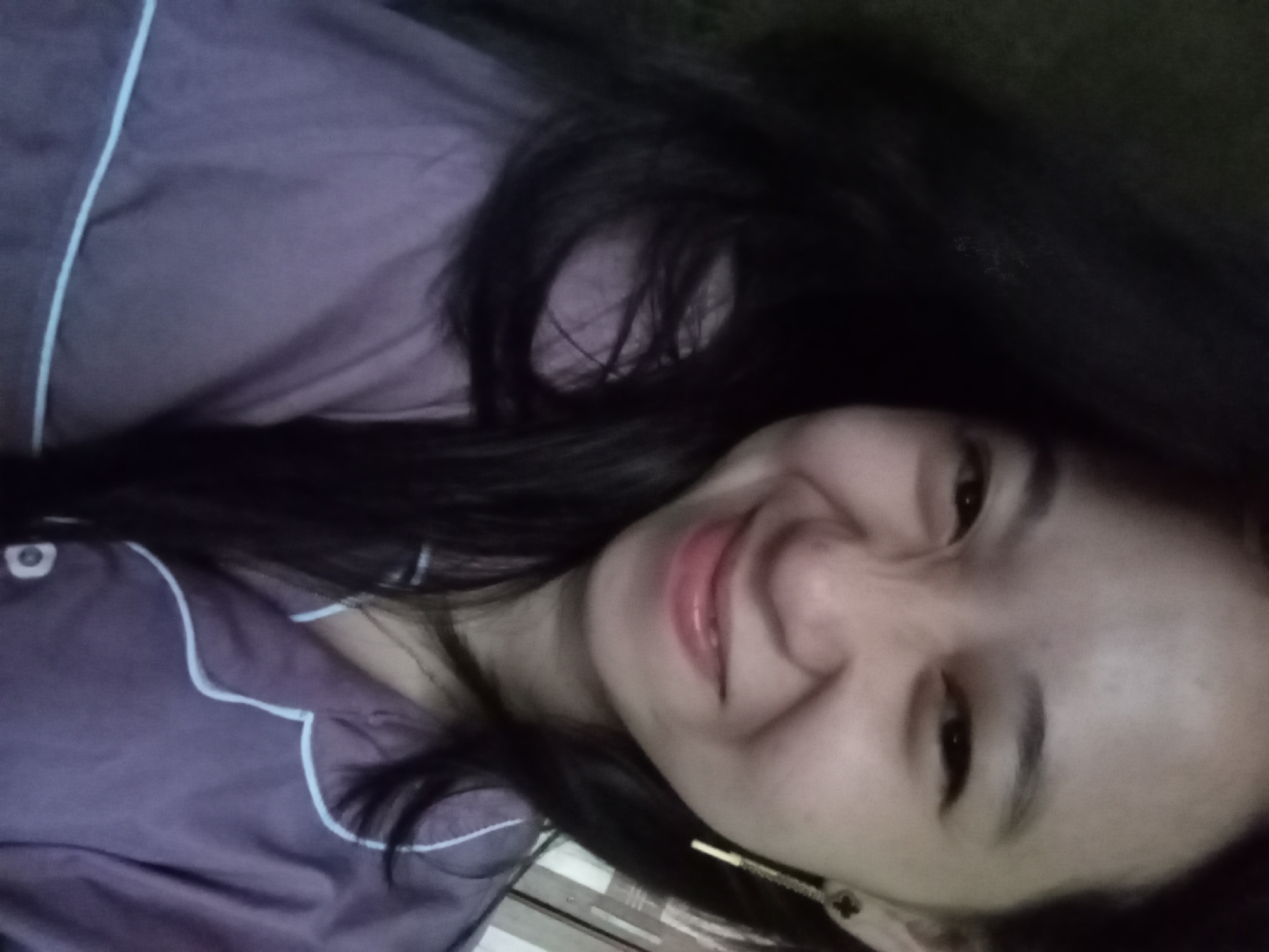 A young woman with long, dark hair is captured in a close-up, selfie-style shot. She is wearing a purple pajama top with white piping. She has a playful expression, with her eyes squinted and a slight smile. A delicate gold earring with a clover-like charm hangs from her right ear. The background is dark and out of focus, with hints of what might be a wall with horizontal lines. The image appears to be taken indoors. The specific location in Rejosari, Indonesia, cannot be determined from the visual information provided.