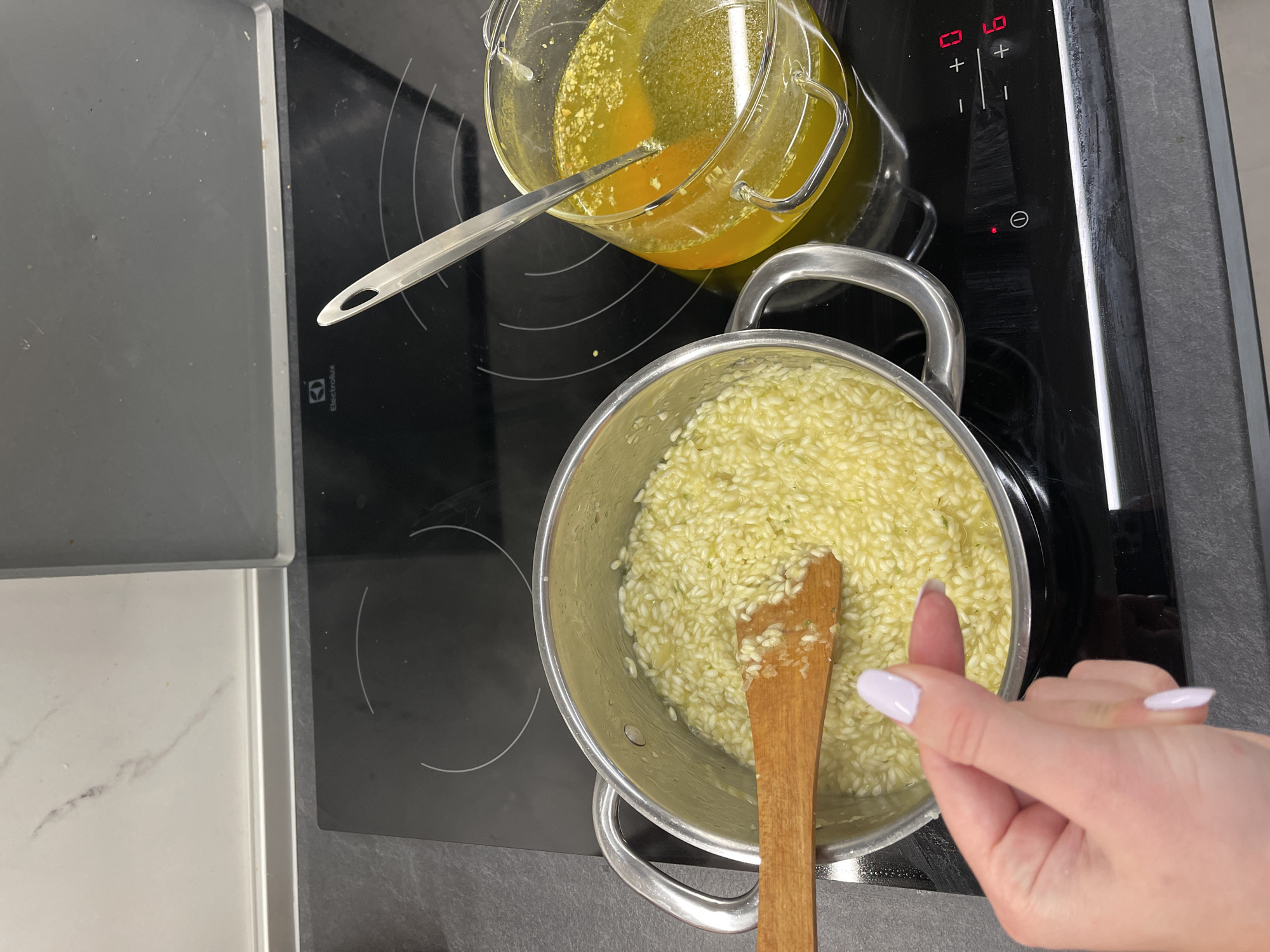 The image shows a cooking scene on a black induction cooktop. A silver pot filled with rice and liquid sits on the cooktop with a wooden spoon inside. Beside the pot is a clear glass container filled with a yellowish liquid. A silver spoon rests inside the container. A hand with light-colored nail polish is reaching toward the pot. The cooktop has touch controls for the heat settings, indicating a modern kitchen setting. The location is not discernible from the visual content of the image.